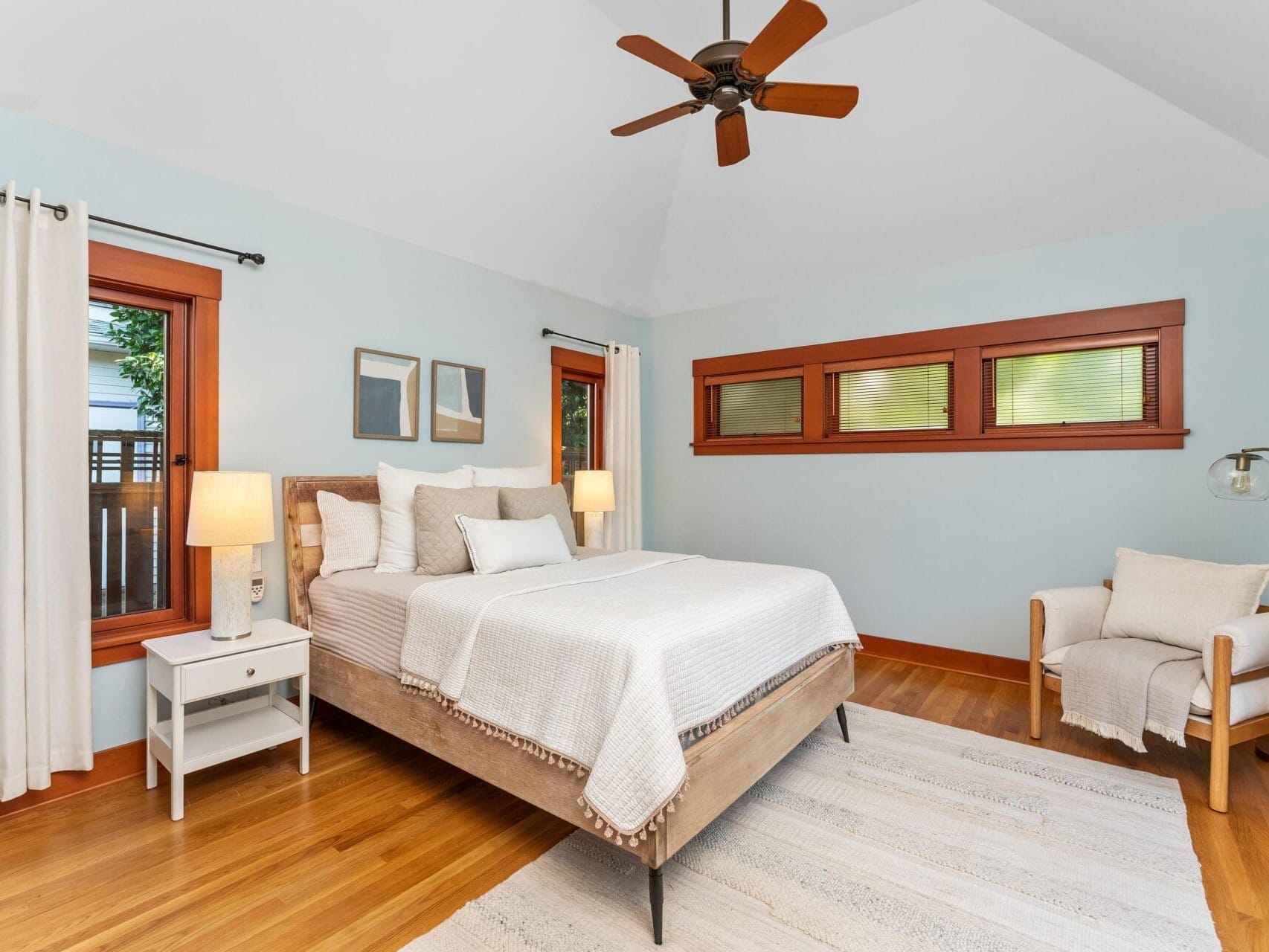 A serene Portland, Oregon bedroom with a large bed featuring white linens and a wooden headboard. There's a side table with a lamp, a cozy armchair, ceiling fan, and two small windows above the bed. Light blue walls and hardwood floors create a calming atmosphere.