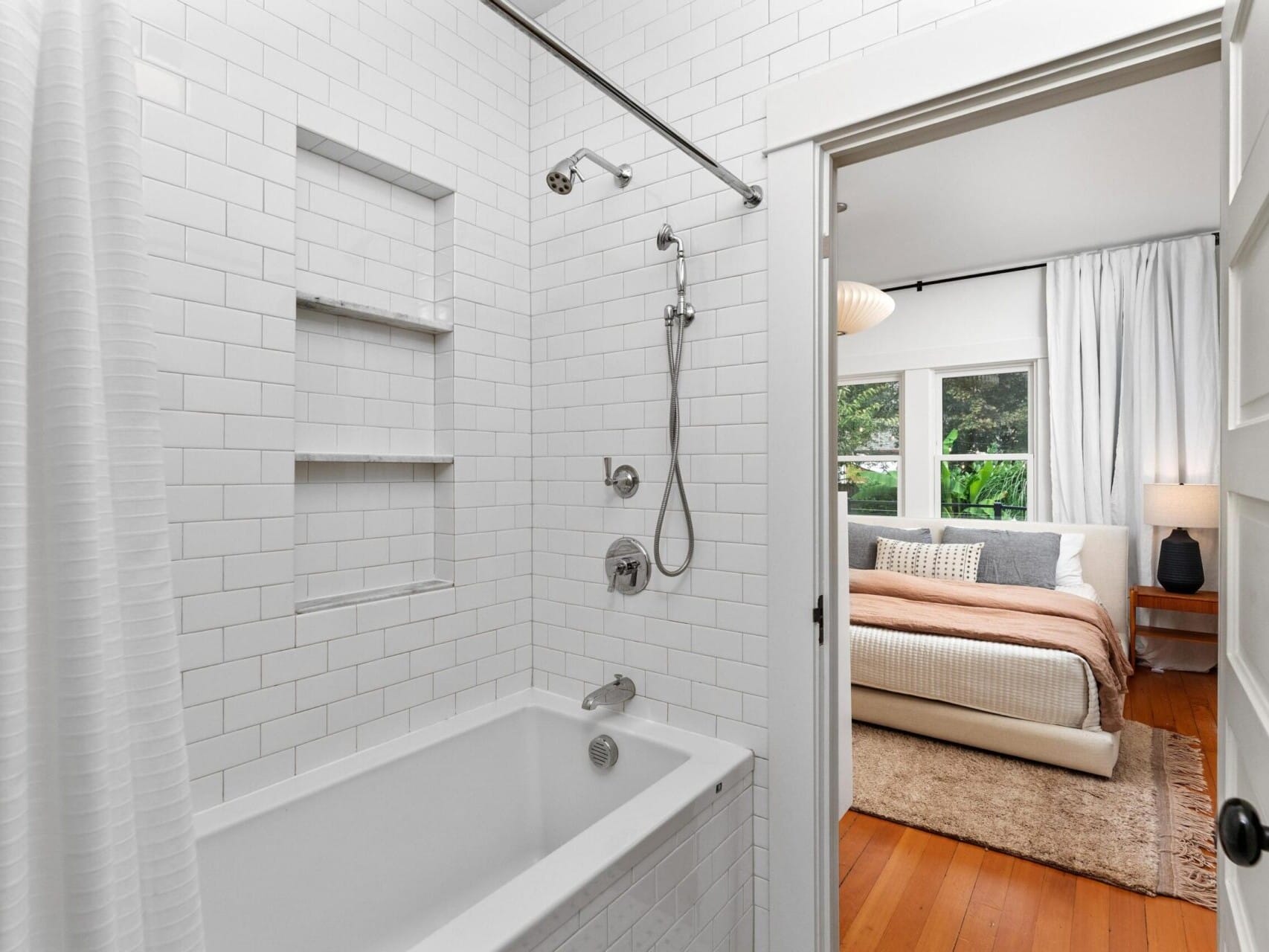 A bathroom with white tiled walls, a bathtub, and a showerhead is visible. This cozy setup in Portland, Oregon connects to a bedroom featuring white and beige bedding, a window with curtains, and a bedside lamp on a wooden floor.