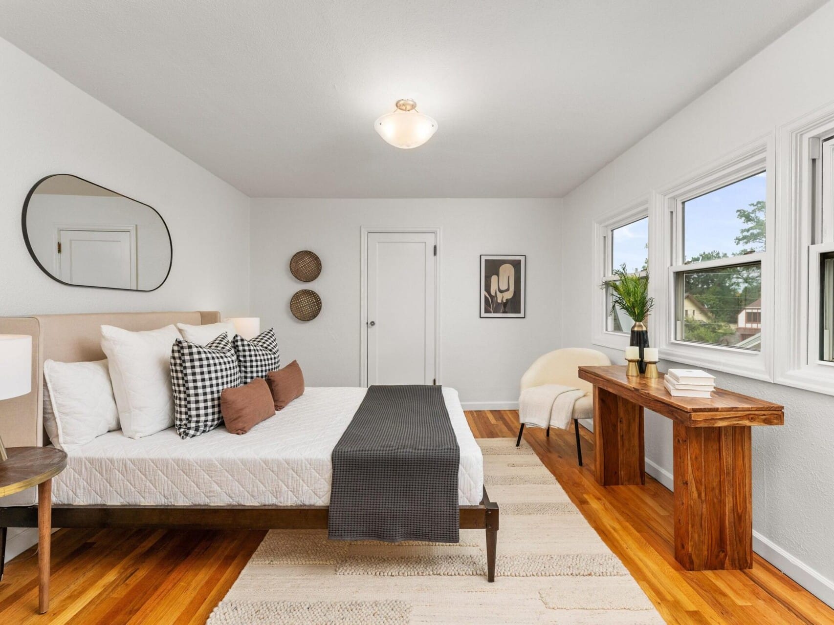 A bright bedroom with a neatly made bed featuring white and patterned pillows, a dark gray throw, and a wooden frame. A large window, a rustic wooden desk, a white chair, and minimal wall art enhance the simple, airy decor.