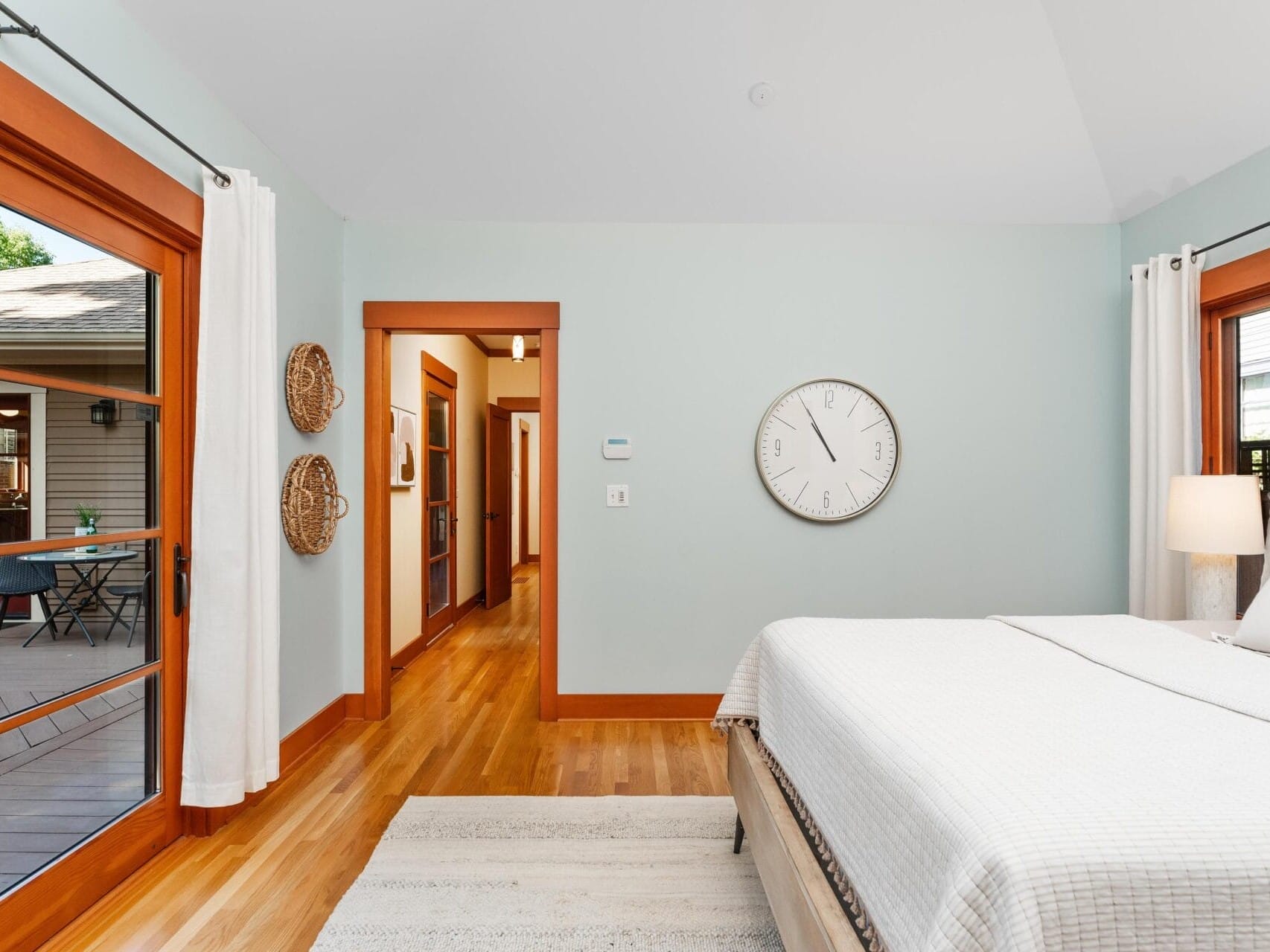 A bright, airy bedroom in Portland, Oregon features a large bed with white linens. Wooden framed glass doors open to an outdoor space. The room boasts light green walls, a large round clock, and two woven wall hangings, all illuminated by natural light.