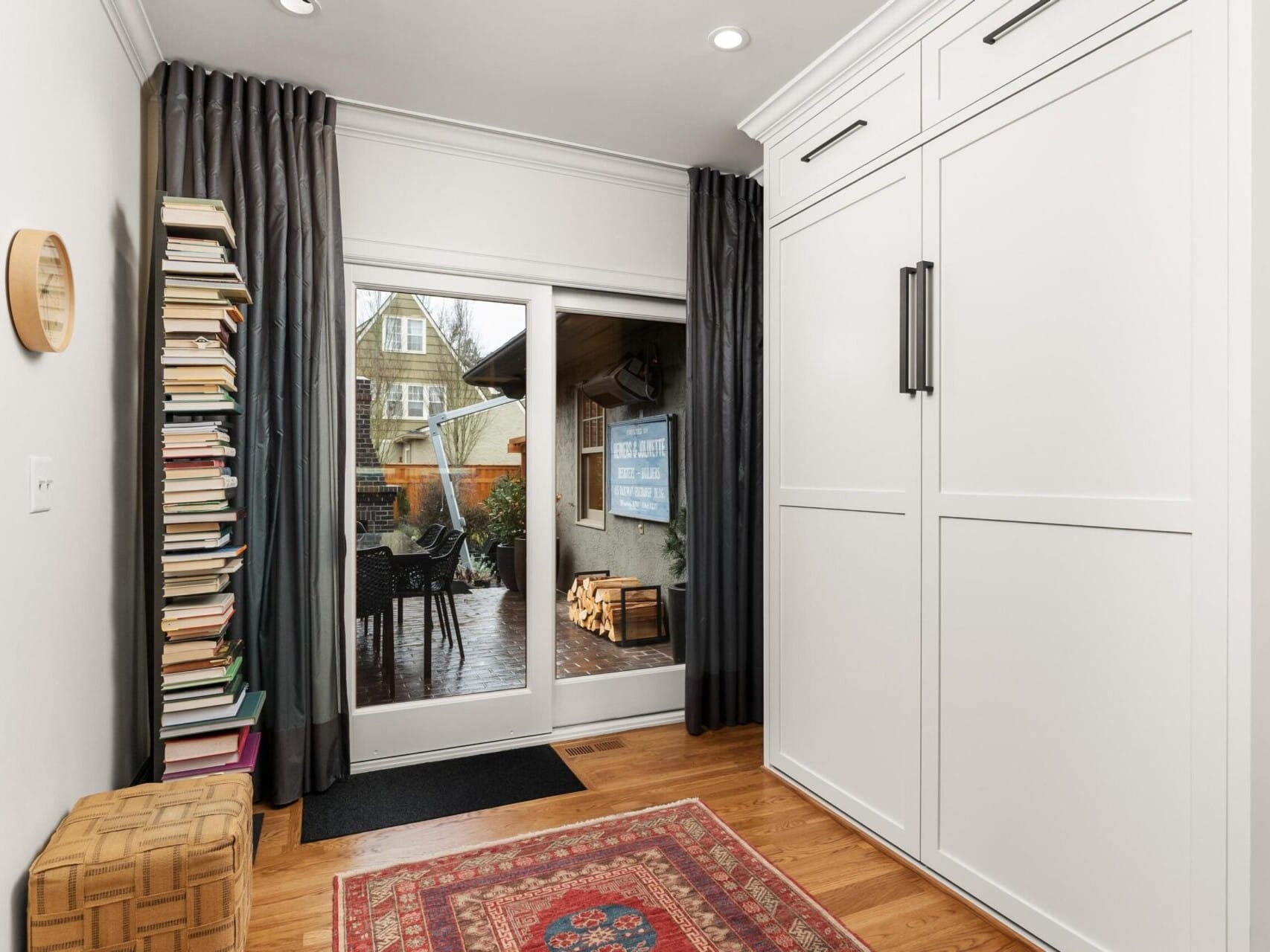 A cozy room with wooden flooring and a colorful rug, perfect for Portland Oregon real estate seekers. A stack of books rests on a shelf to the left, while sliding glass doors lead to a patio with chairs and views of neighboring houses. Large white cabinets stand to the right, framed by elegant curtains.