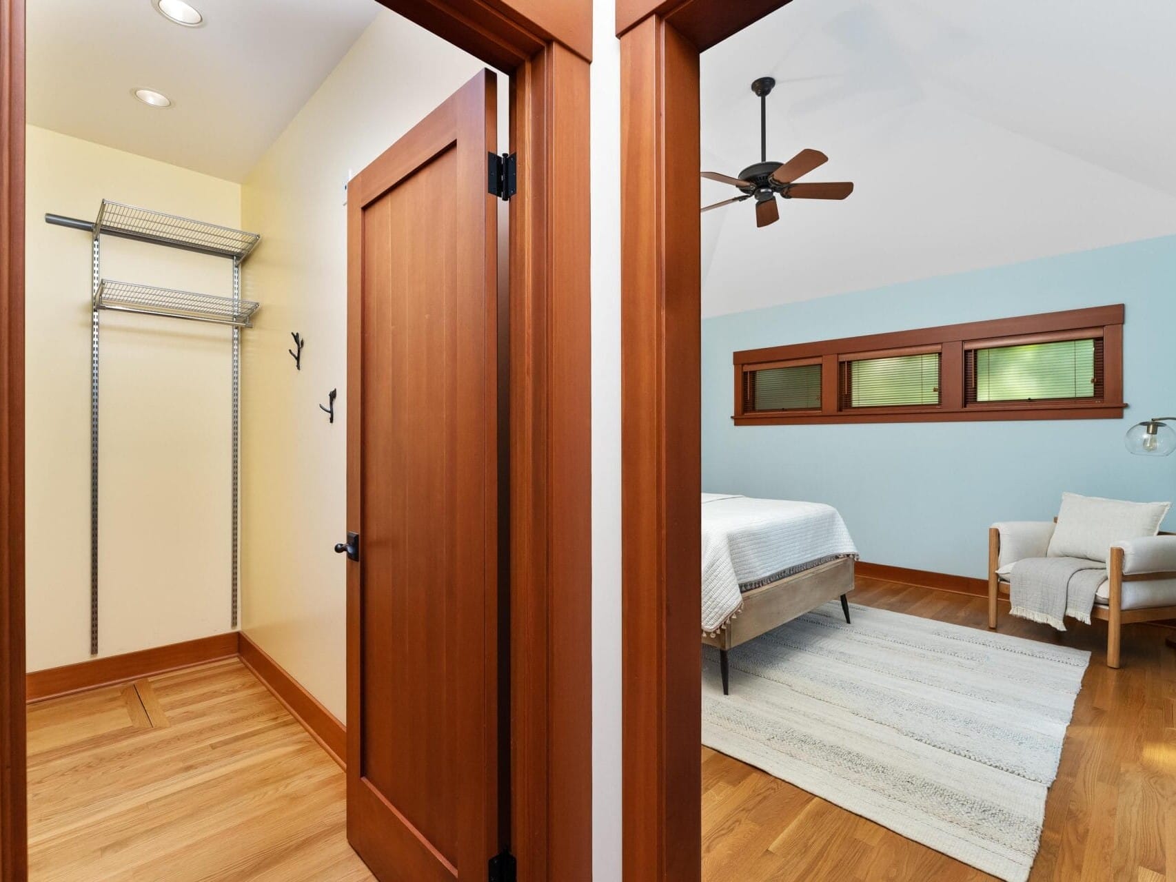 A hallway with a view into a bedroom on the right and an empty closet area on the left. The Portland, Oregon bedroom features a bed against a light blue accent wall, wooden floor, ceiling fan, cozy chair draped with a blanket, and a window with three panes.