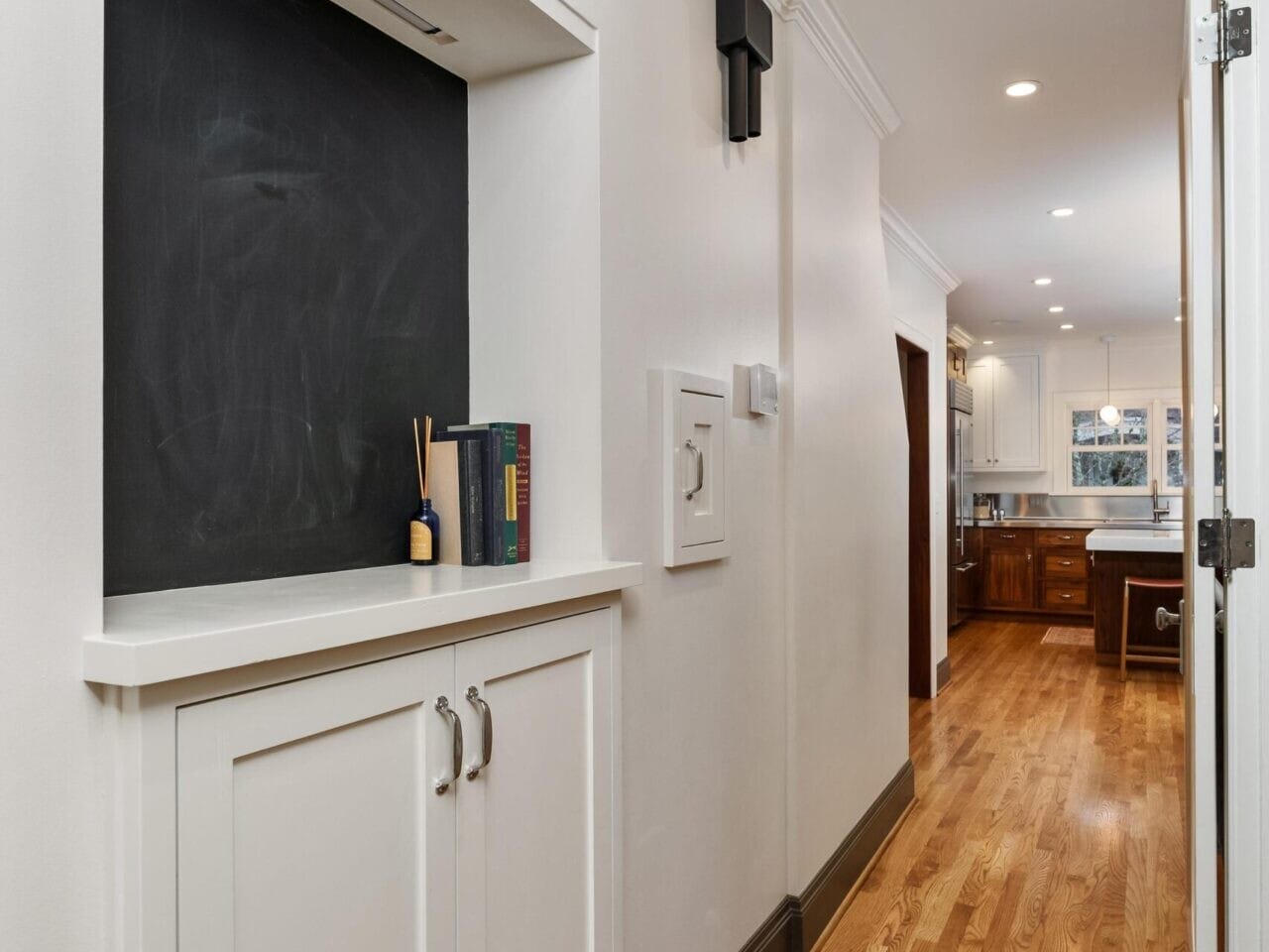 A hallway with wooden flooring leads to a kitchen. On the left, there’s a chalkboard nook above a white cabinet with a reed diffuser and books. The walls are white, and the space is lit with recessed lighting—typical of Portland Oregon real estate's charming interiors.