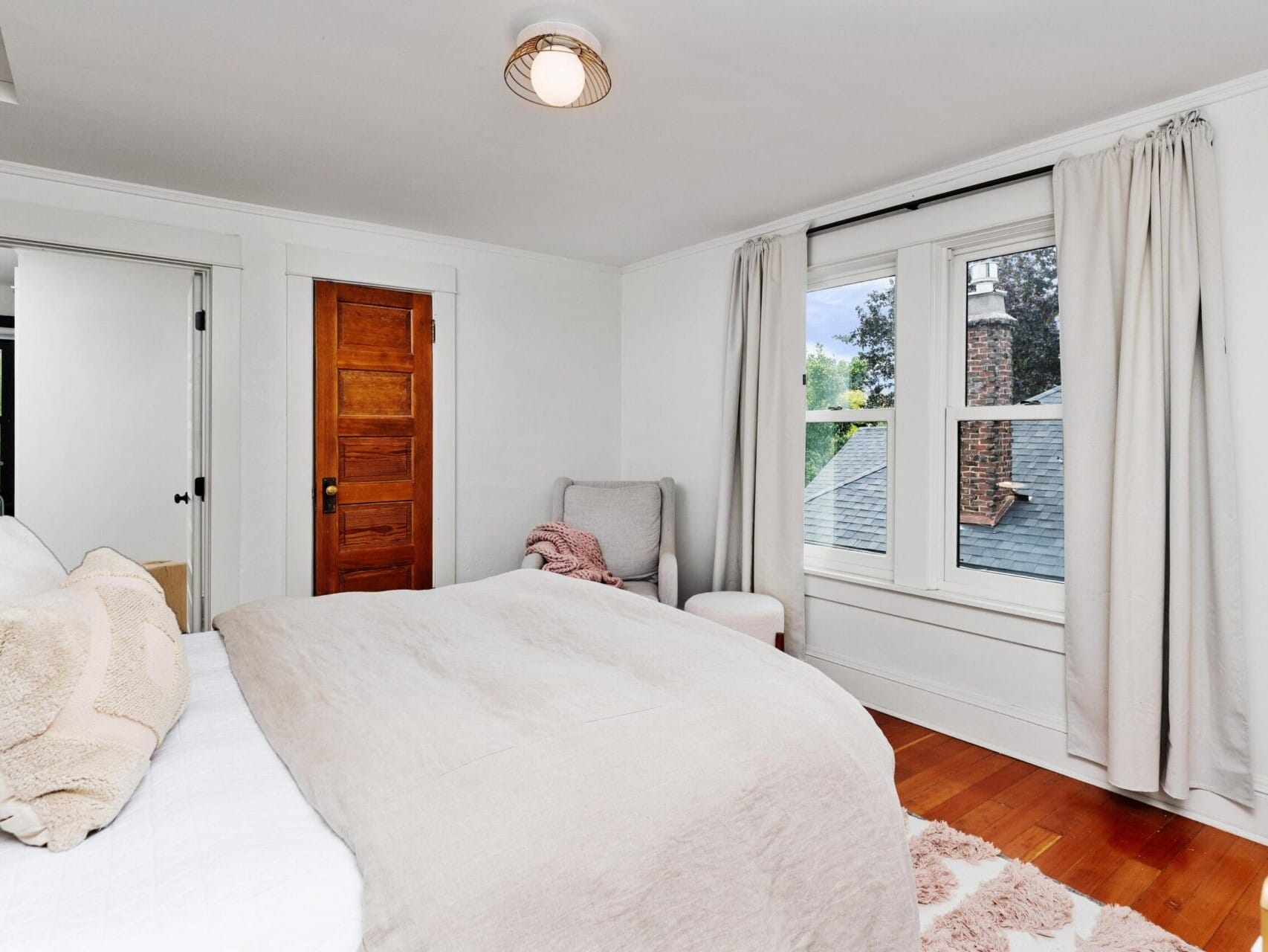 Cozy Portland bedroom with a large bed, cushioned with pillows and a beige comforter. A wooden door stands in the corner, flanked by a window with curtains. A soft armchair and ottoman sit beside the window, enhancing the serene ambiance typical of Oregon charm.