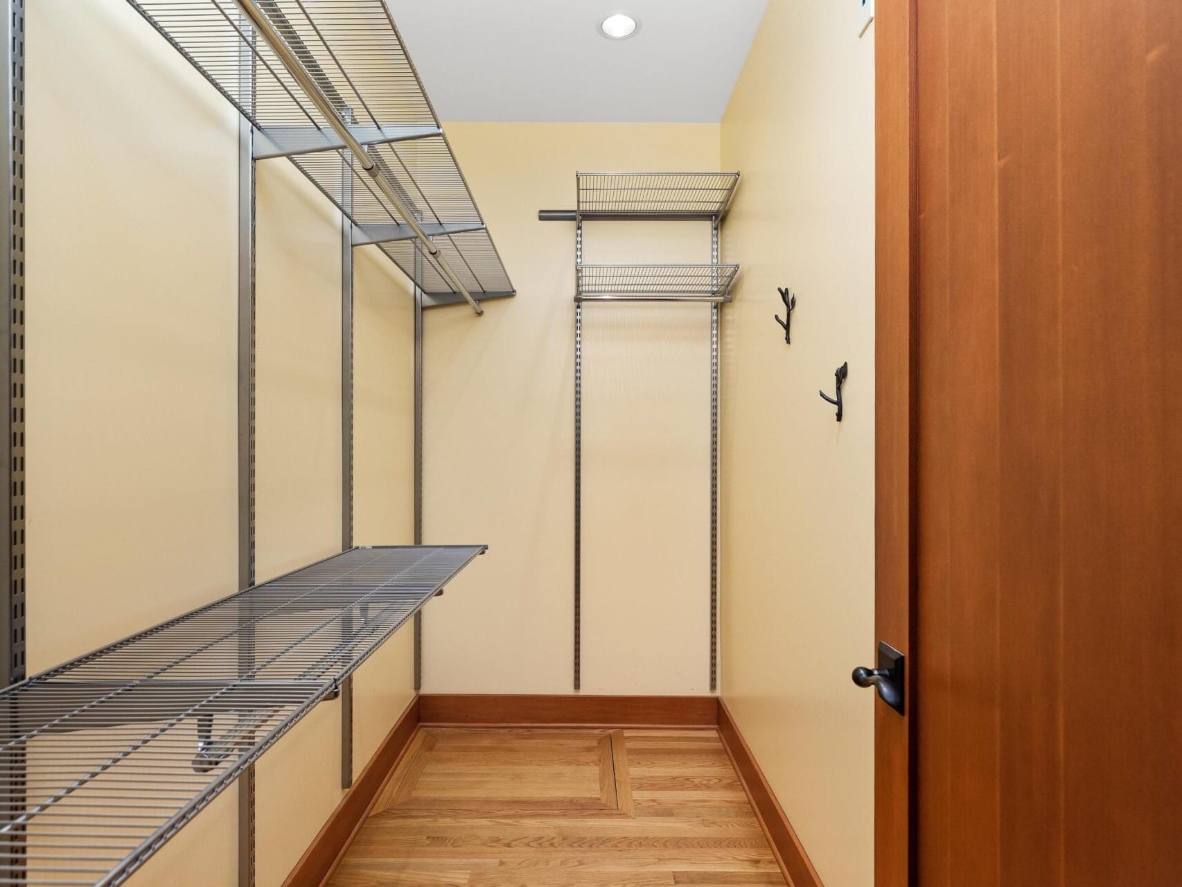 An empty walk-in closet in a Portland, Oregon home showcases beige walls and metal wire shelving on two sides. The hardwood floor complements the wooden door adorned with black metal hooks, while ceiling lights provide ample illumination.