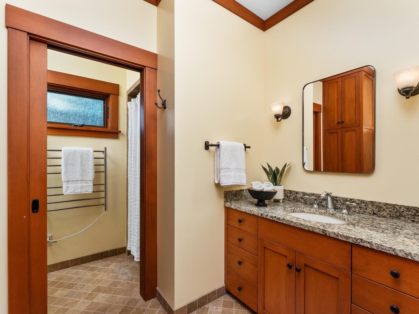 A bathroom in Portland, Oregon, features a wooden cabinet vanity with a granite countertop. Above the sink, a large mirror and wall-mounted light fixture add elegance. The room's tan walls and brown tile flooring complement the shower, visible through an open door with a white curtain.