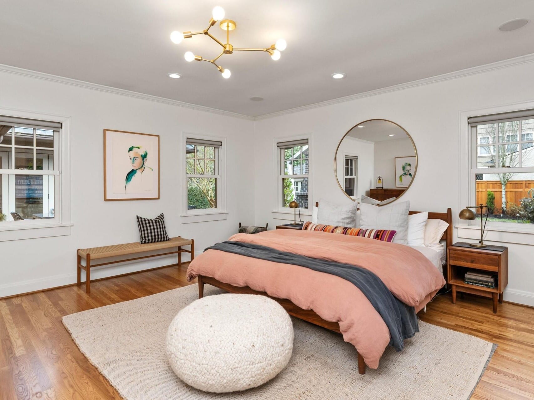 A contemporary bedroom in a Portland Oregon real estate gem features a wooden bed with peach and gray bedding, a large round mirror, and a modern chandelier. A bench sits under a wall photo, and a knitted pouf rests on a light rug. Multiple windows bring in natural light.