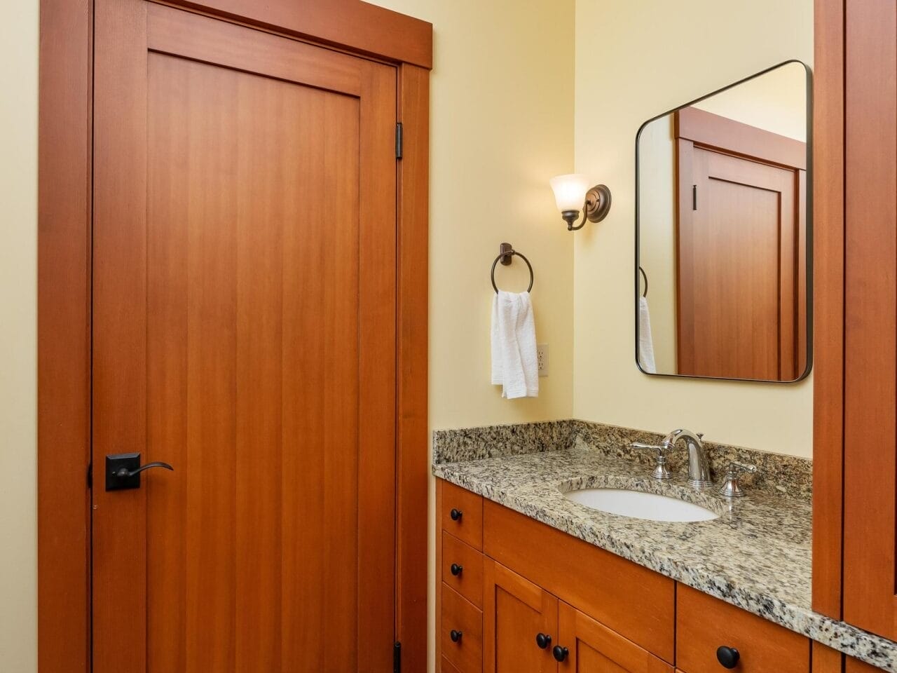 This Portland, Oregon bathroom boasts a wooden door and cabinetry, paired with a granite countertop and sink. A rectangular mirror adorns the wall above the sink, accompanied by a nearby towel ring. The floor showcases an elegant beige and brown tile pattern.