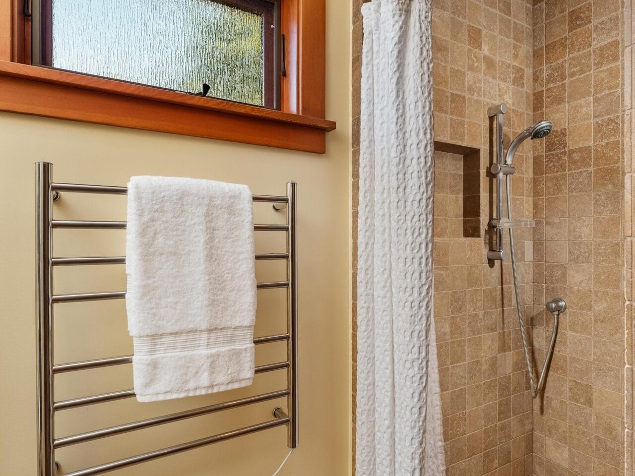 A bathroom in Portland, Oregon, features a beige tiled walk-in shower with a crisp white curtain. A towel hangs neatly on the heated rail to the left, while a high frosted window above adds an airy touch.