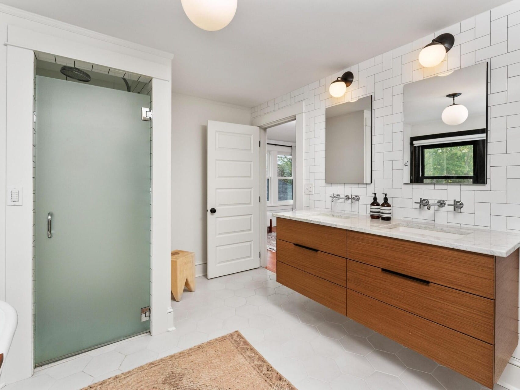 Modern bathroom in Portland, Oregon, with white hexagonal tile flooring and a double vanity featuring wooden drawers and white countertops. Two mirrors and lights grace the sinks, while a frosted glass shower is on the left. A wooden stool complements the cozy rug on the floor.