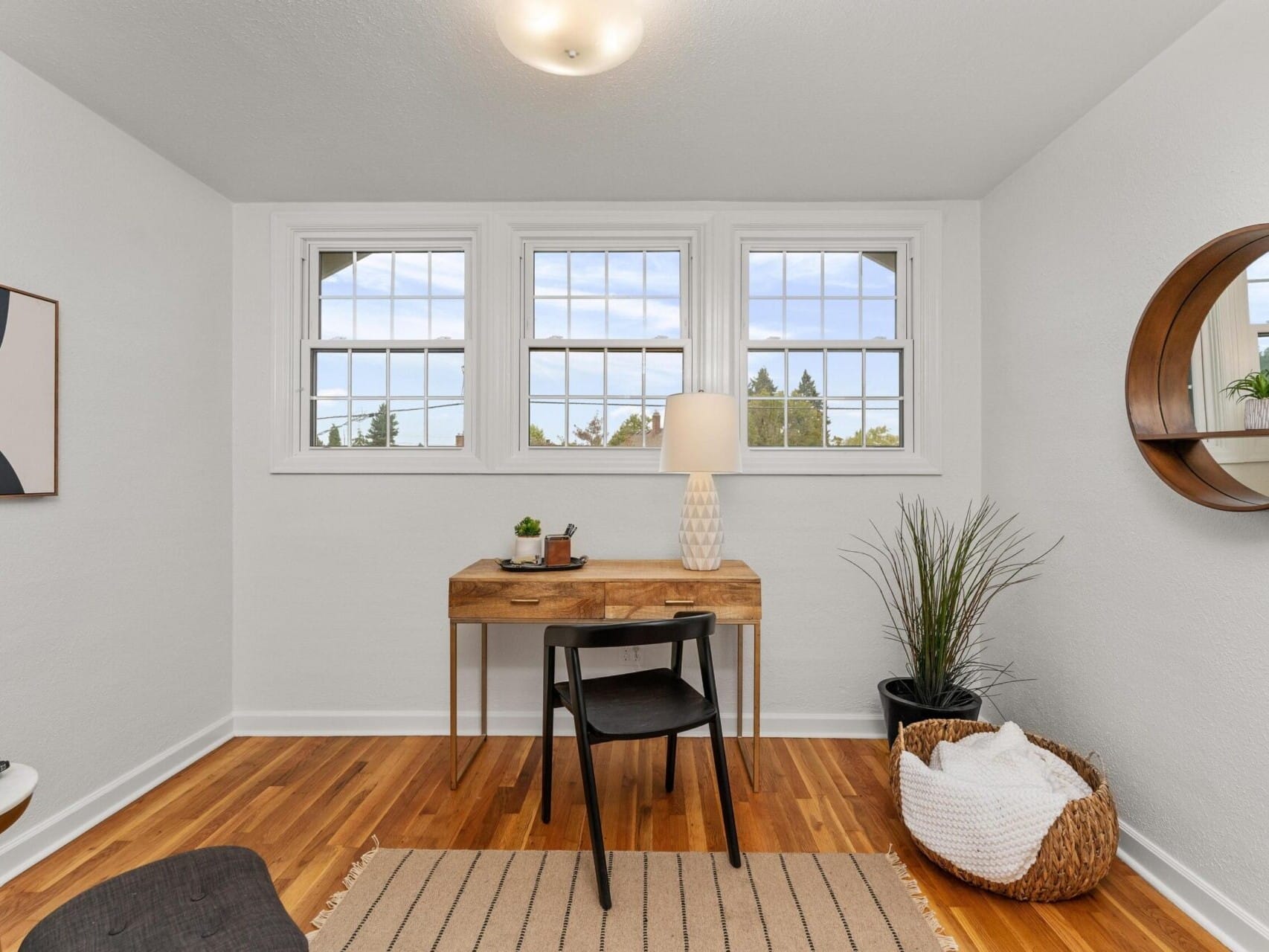 A small, minimalist home office with a wooden desk and black chair. The desk holds a lamp and plant. Three windows provide natural light. A round mirror and a basket with a blanket are beside the desk. White walls and wooden floor create a cozy feel.