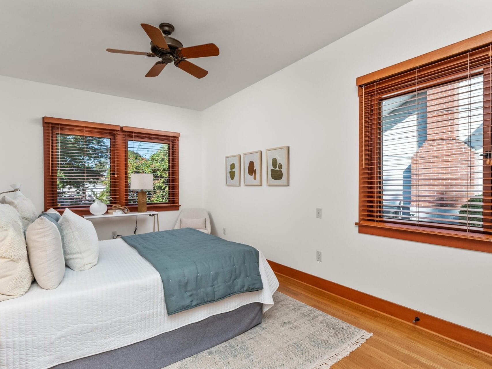 A cozy Portland, Oregon bedroom features a white bed adorned with a blue throw and several pillows. Wooden blinds filter natural light through windows on two walls. A small desk with a lamp sits by the window, under three abstract artworks. Overhead, a ceiling fan gently whirs.