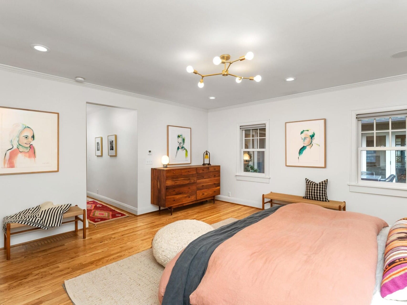 A modern bedroom with wooden flooring features a large bed with a peach duvet. There's a wooden dresser, a bench, and four colorful abstract portraits on the walls. A unique ceiling light fixture adds a contemporary touch. Two windows brighten the room, capturing that Portland Oregon real estate charm.