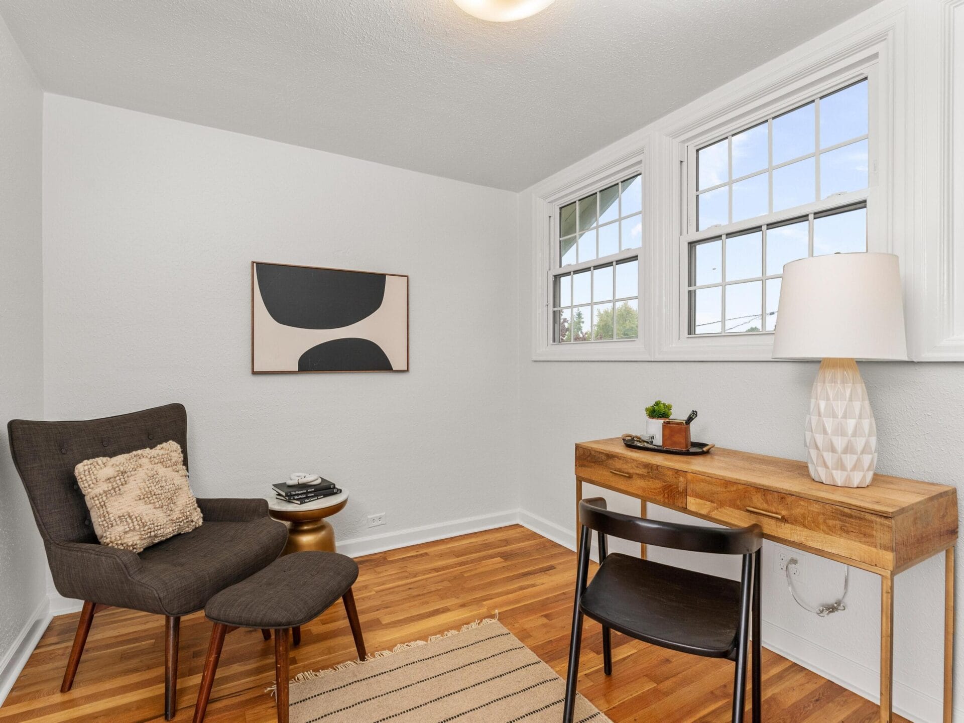 A cozy room with a dark armchair and ottoman, a small side table with books, a wooden desk with a chair, and a lamp. A modern abstract painting hangs on the wall, and two windows allow natural light to fill the space. A beige rug lies on the hardwood floor.