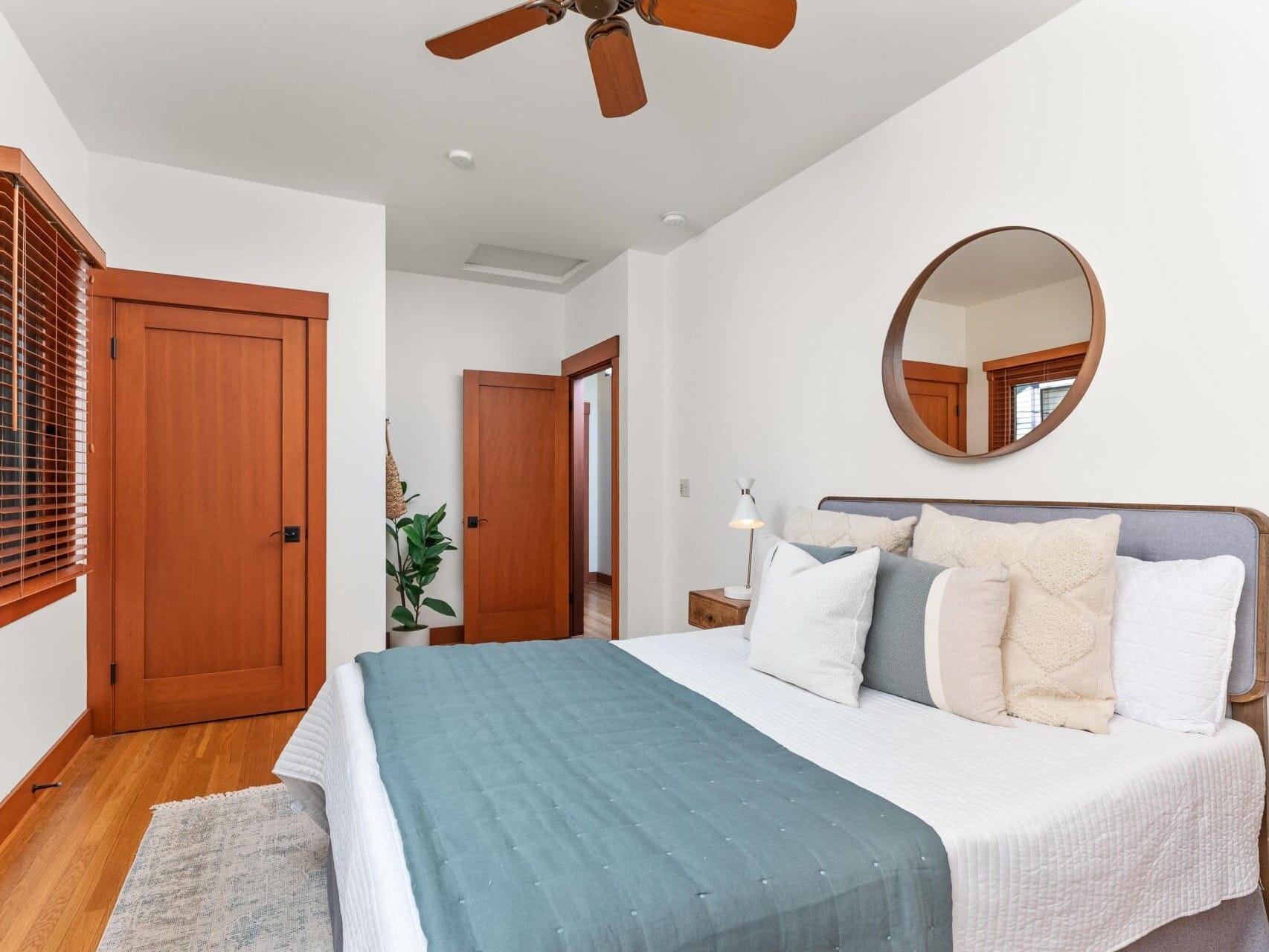 A cozy Portland, Oregon bedroom with a neatly made bed featuring white and green bedding and multiple pillows. A round mirror hangs above the bed, accompanied by a wooden side table with a lamp. A ceiling fan is overhead, and the room boasts wooden doors and window frames.