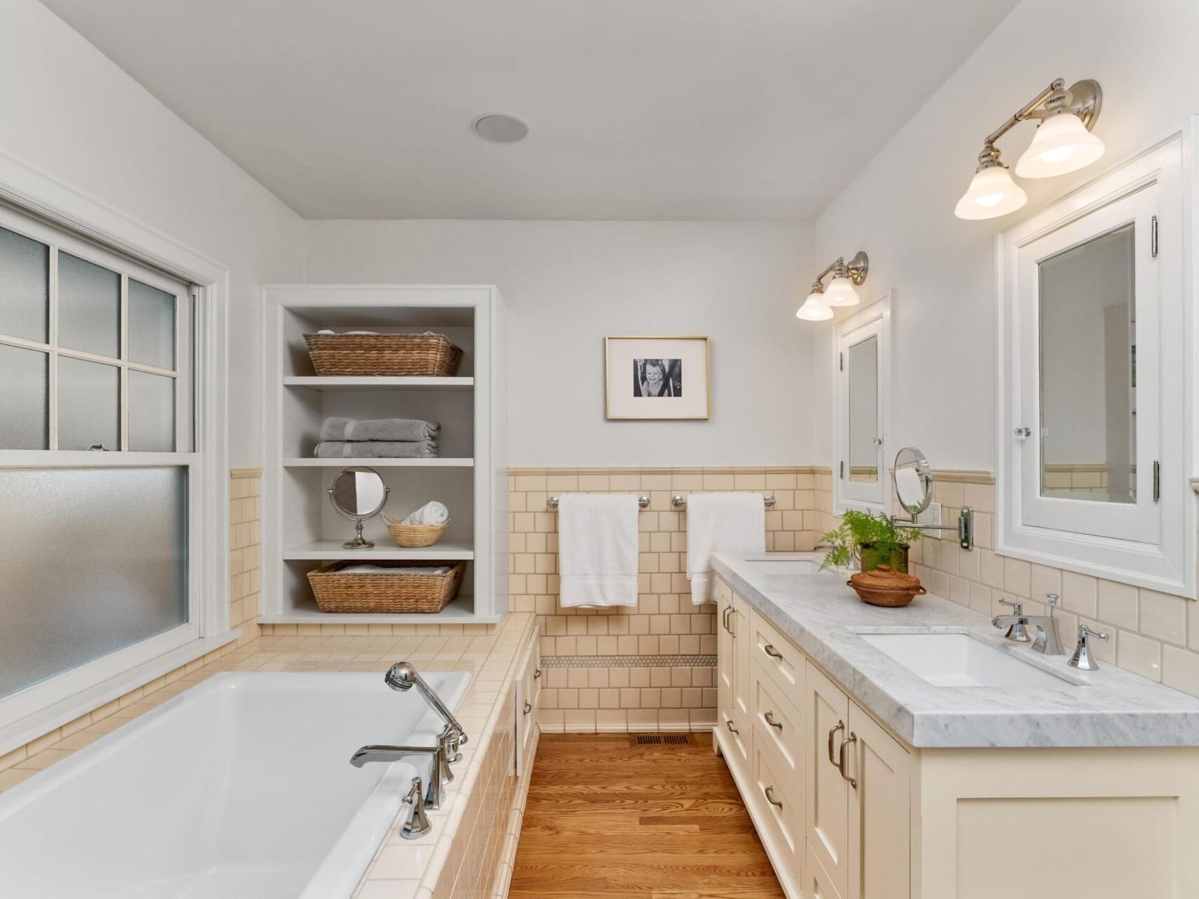 A bright bathroom in a top Portland Oregon real estate listing features a window, built-in shelving, and a bathtub on the left. On the right, there's a double sink vanity with a marble countertop, decorative lighting, and wooden flooring. Neutral tones create a serene atmosphere.