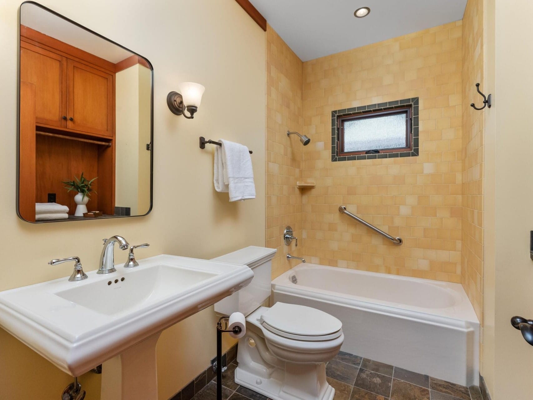 A charming bathroom in Portland, Oregon features a pedestal sink, mirror, and light fixture on the left. The right side boasts a bathtub with beige tiled walls and a window. Complete with a toilet and towel rack holding a neatly folded towel above it, this space is both functional and inviting.