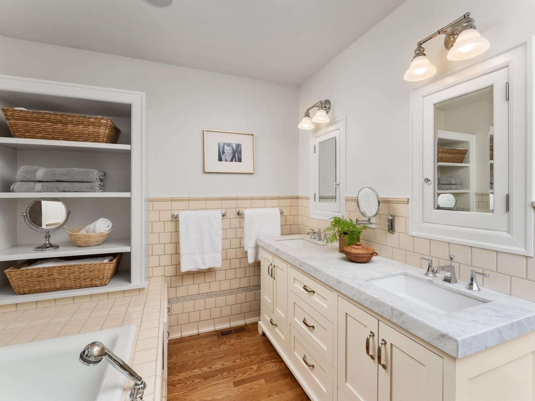 A bright bathroom, perfect for a Portland Oregon Real Estate listing, featuring a double sink vanity with a marble countertop and silver faucets. Above are two light fixtures. Towels hang on the wall, while shelves with wicker baskets and folded towels are visible. A plant adds a touch of greenery.