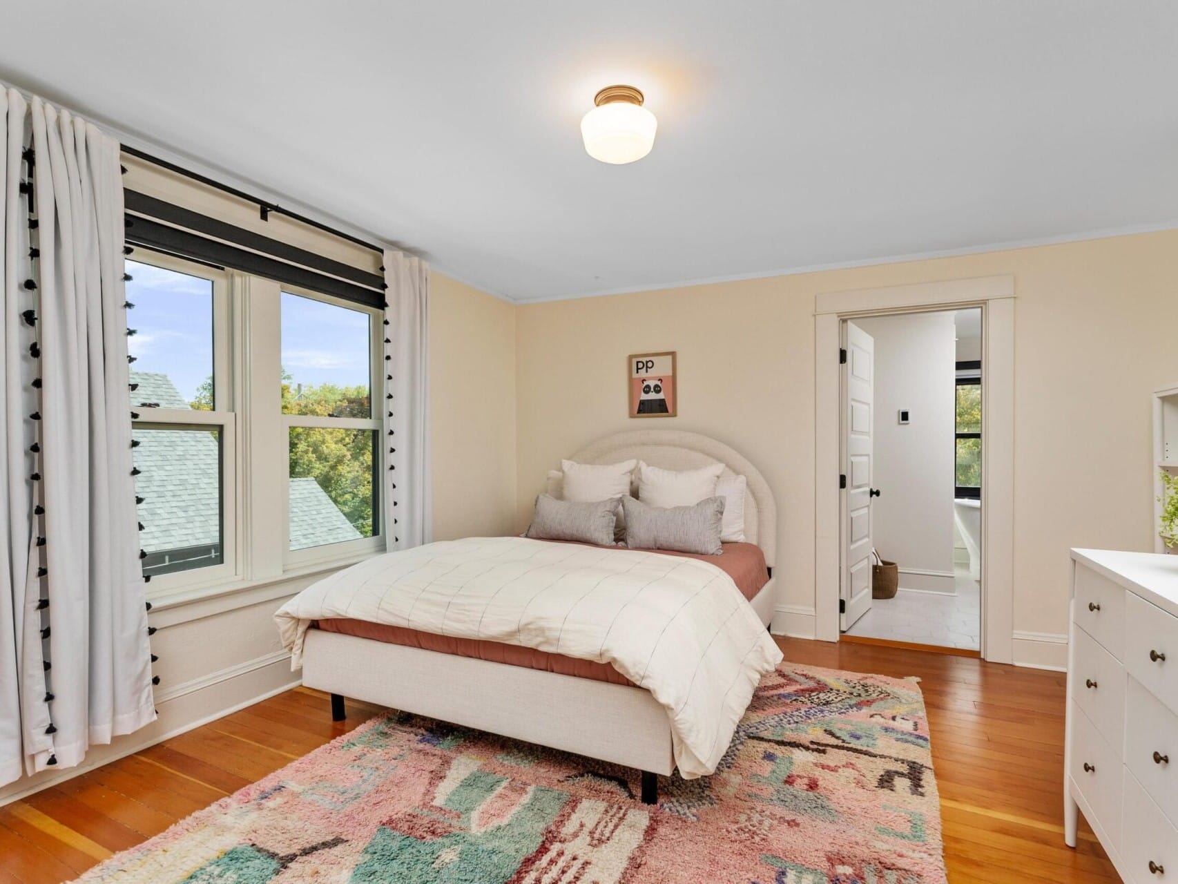 A cozy Portland, Oregon bedroom with a neatly made bed, light-colored walls, and wooden floors. A colorful rug lies under the bed. The room features a window with curtains, a dresser adorned with plants, and an open door leading to another inviting space.