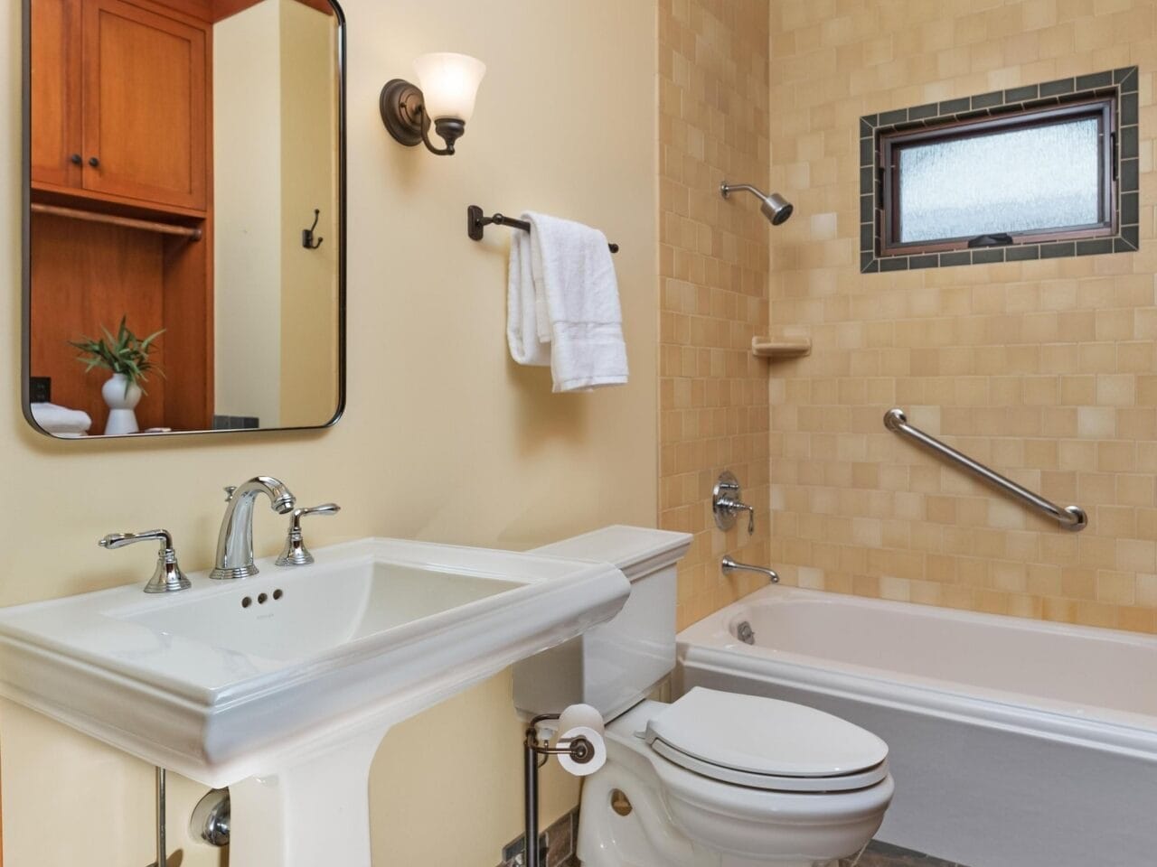 This Portland, Oregon bathroom features a pedestal sink, toilet, and bathtub with a grab bar. It includes a wall-mounted mirror, sconce lighting, and beige tiles. The floor is brown stone, and there's a small window above the bathtub with a towel hanging nearby.