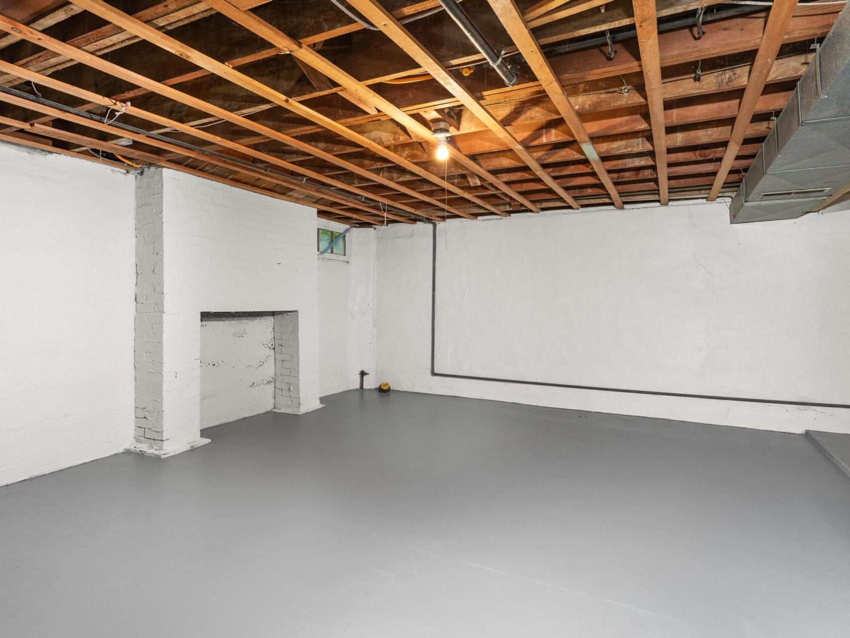 A spacious basement with painted white walls and exposed wooden beams on the ceiling. The floor is a smooth, light gray. A small window lets in some natural light, and theres a light bulb hanging from the ceiling.