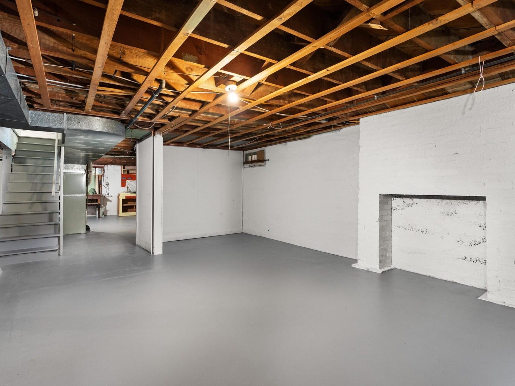 Spacious basement with exposed wooden beams, white painted brick fireplace, and concrete floors. Open layout with a staircase at one end and unfinished walls, giving an industrial look.