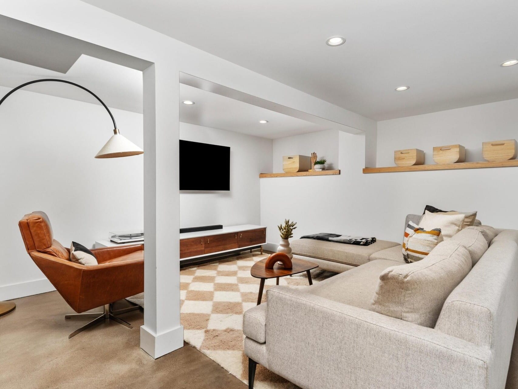 Modern living room in a Portland, Oregon home with a beige sectional sofa, brown leather armchair, and floor lamp. A wall-mounted TV is opposite the seating area. Wooden shelves with decorative boxes are mounted on the far wall. A geometric rug covers the floor.