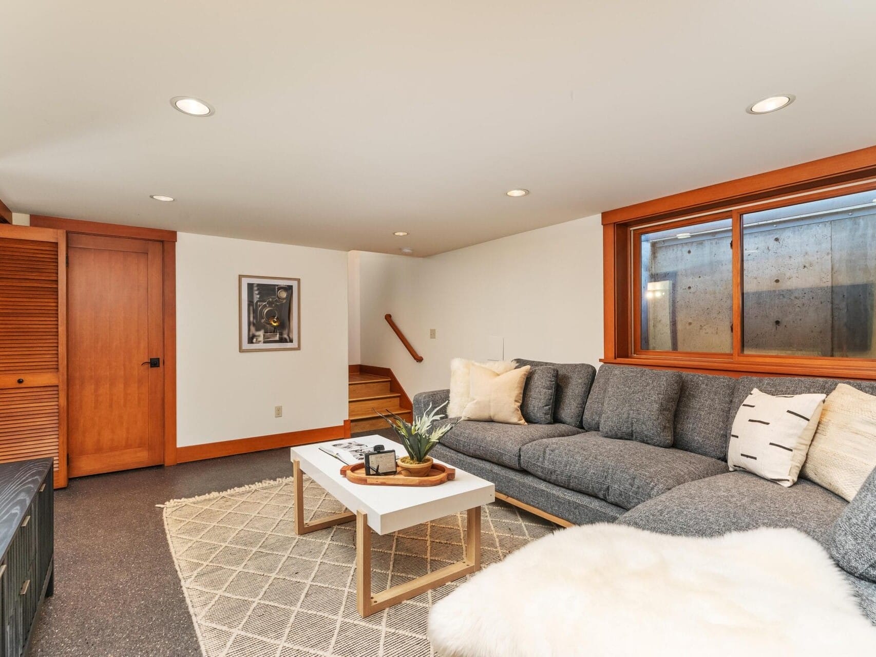 A cozy living room in Portland, Oregon, featuring a gray sectional sofa with white pillows and a fluffy throw. A wooden coffee table rests on a patterned rug. A wooden door and staircase lead upstairs, while a large window illuminates the space, complemented by framed artwork on the wall.