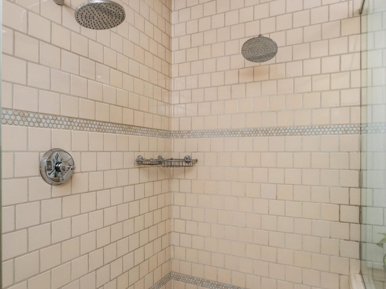 A tiled shower in a Portland, Oregon real estate listing features beige and small patterned tiles, two overhead showerheads, a convenient glass shelf for toiletries, and a sleek glass partition on the right side. Perfectly presented by your top real estate agent.