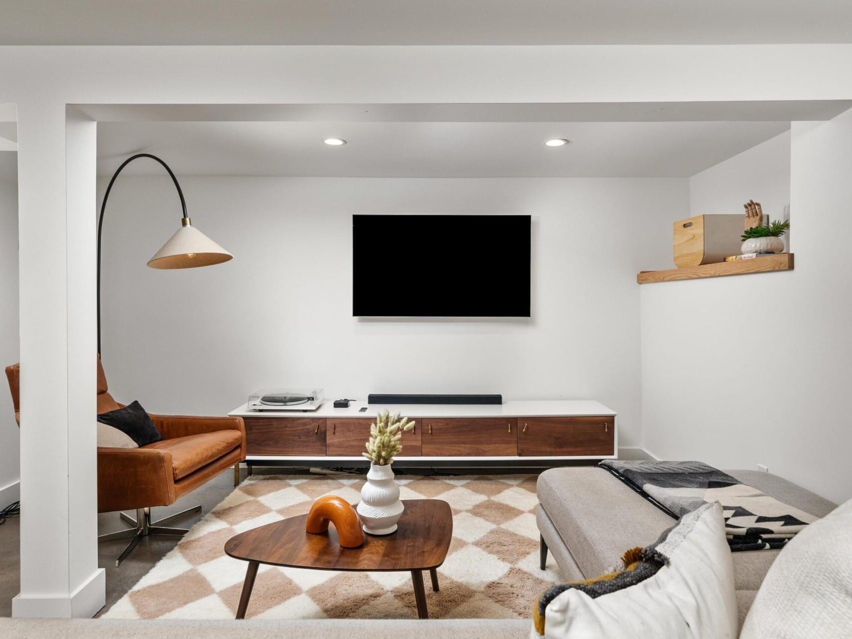 In a modern Portland, Oregon living room, a wall-mounted TV complements a sleek wooden media console. A cozy seating area features a brown leather chair and beige sofa, with a stylish floor lamp arching overhead. Decorative items are thoughtfully arranged on a floating shelf.