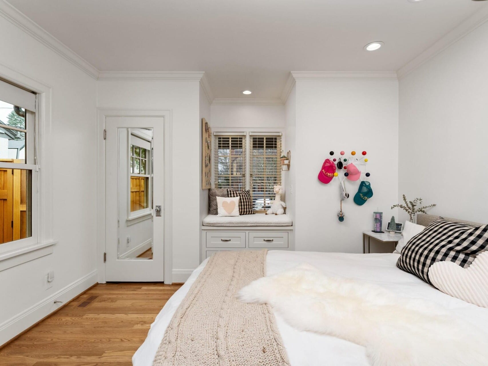 A cozy bedroom in Portland, Oregon real estate features a soft bed adorned with pillows and a knitted throw. A window seat with plush toys sits under a large window, while a wall pegboard holds hats. The room boasts wooden floors and white walls, offering charm and comfort for home seekers.