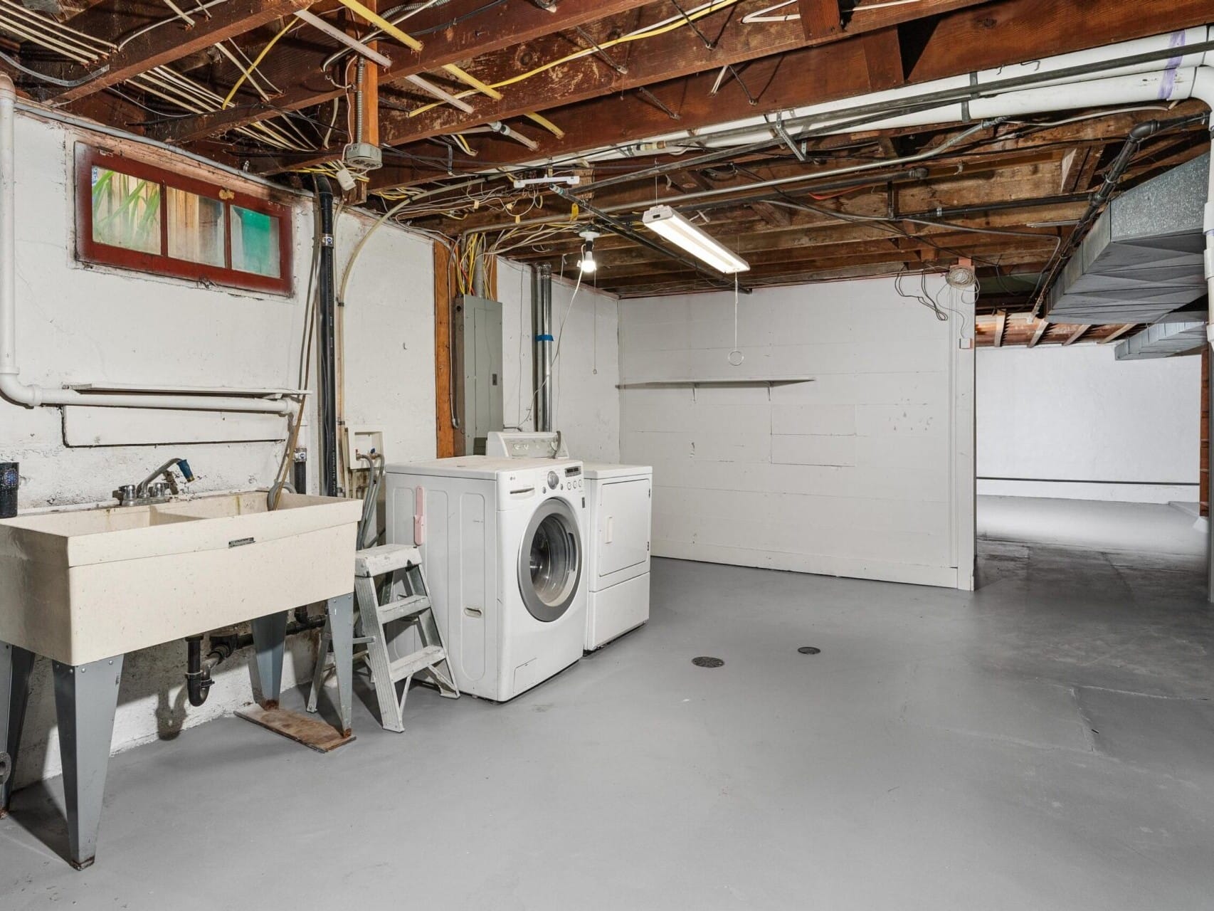 A basement laundry room with a washer, sink, and exposed pipes and beams. The space has concrete floors and walls with a small window, and a step stool is next to the washer. Theres a divider in the back separating another area.