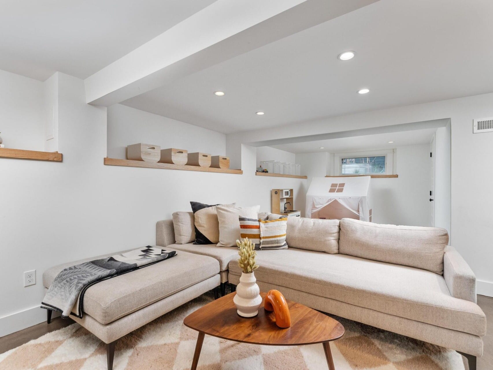 A bright, modern living room in Portland, Oregon, boasts a beige sectional sofa, a patterned rug, and a triangular wooden coffee table. The space features recessed lighting, wall-mounted shelves with baskets, and a child's play area complete with a small tent in the background.