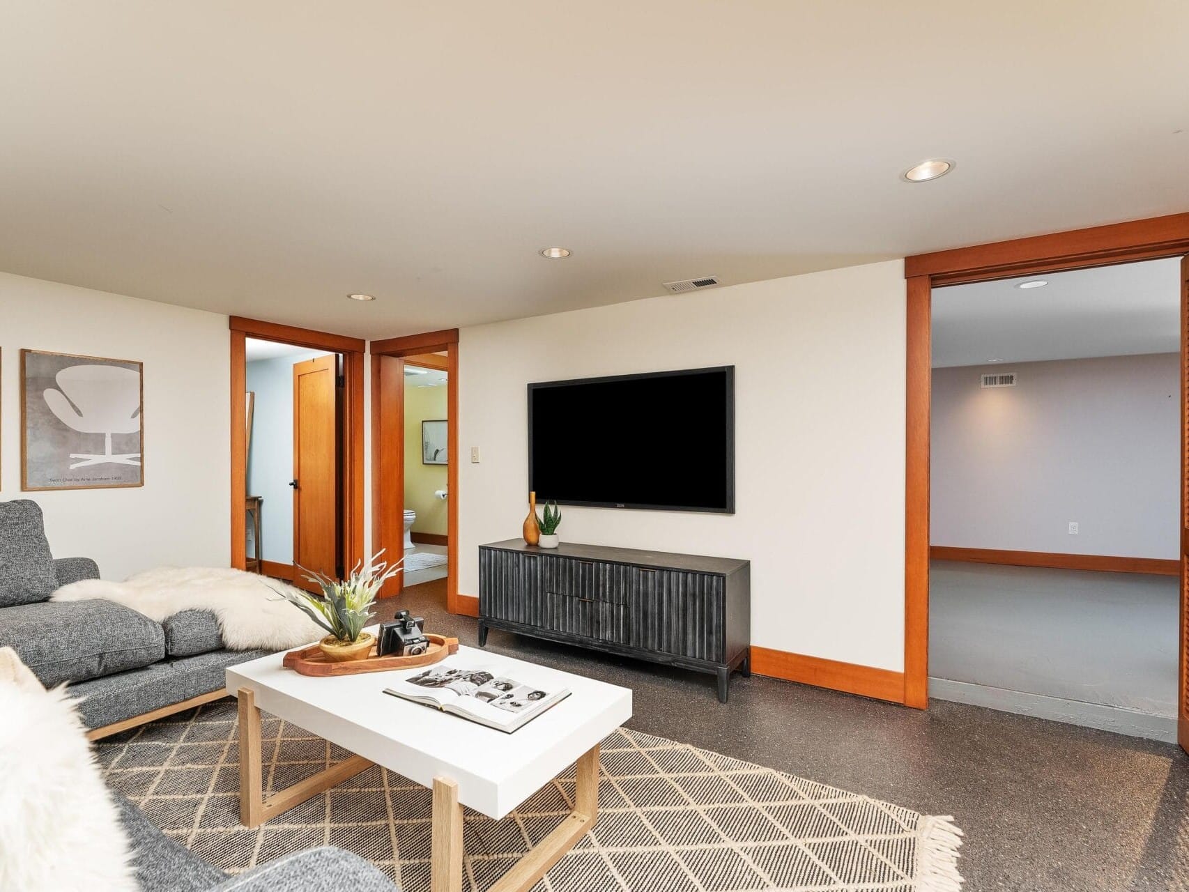 A cozy living room in Portland, Oregon features a gray sectional sofa, fluffy white throw, and wooden coffee table. A large flat-screen TV is mounted on the wall above a black console. The room has beige walls, recessed lighting, and a patterned rug on the floor.