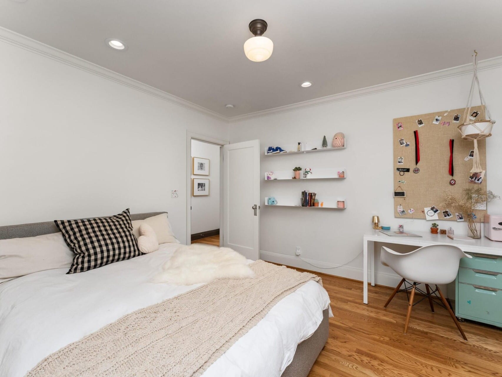 A cozy bedroom with a neatly made bed featuring white and plaid pillows. A study desk with a white chair and pinboard is on the right. Shelves with decor items hang above the desk. The room, showcasing Portland Oregon Real Estate, has wooden flooring and white walls.