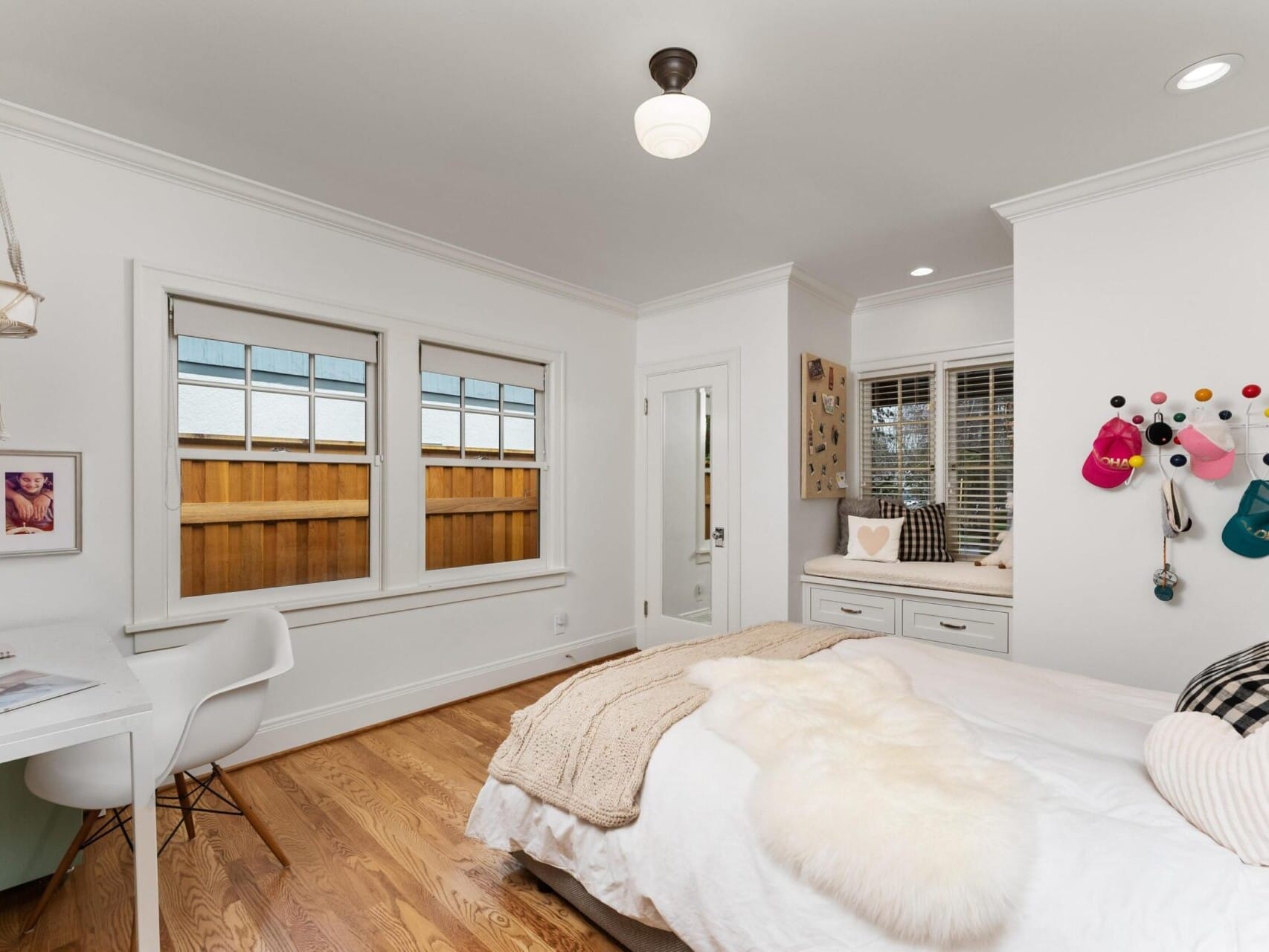 A cozy bedroom in a Portland, Oregon real estate gem features a neatly made bed with a fur throw, a white desk and chair by the window, and colorful hats adorning the wall. Light wood flooring and a window seat with storage underneath complete this inviting space.