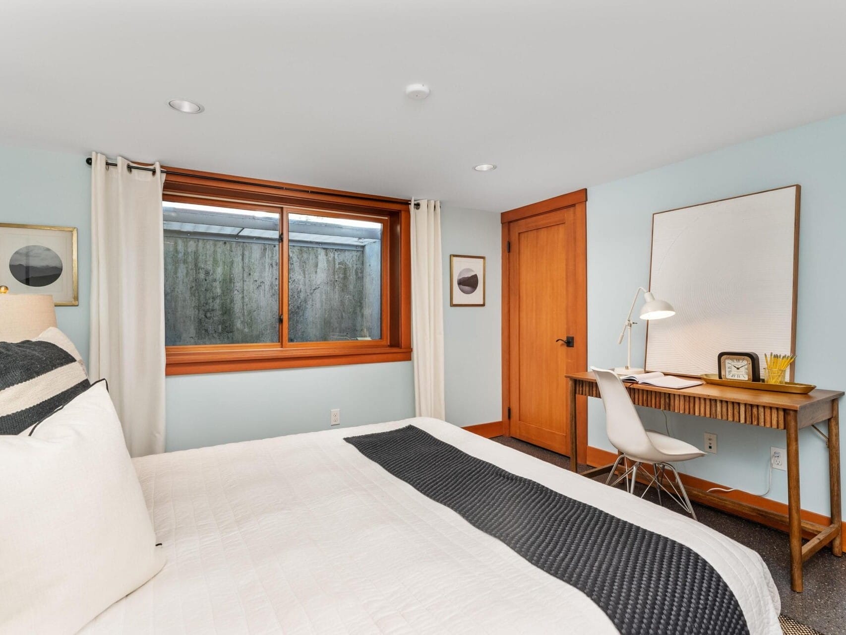 A cozy bedroom in Portland, Oregon, features a large bed with white and black bedding. There's a wooden desk with a lamp and chair against the wall. Soft lighting from a wide window with curtains illuminates the room, while minimalist framed art decorates the light blue walls.