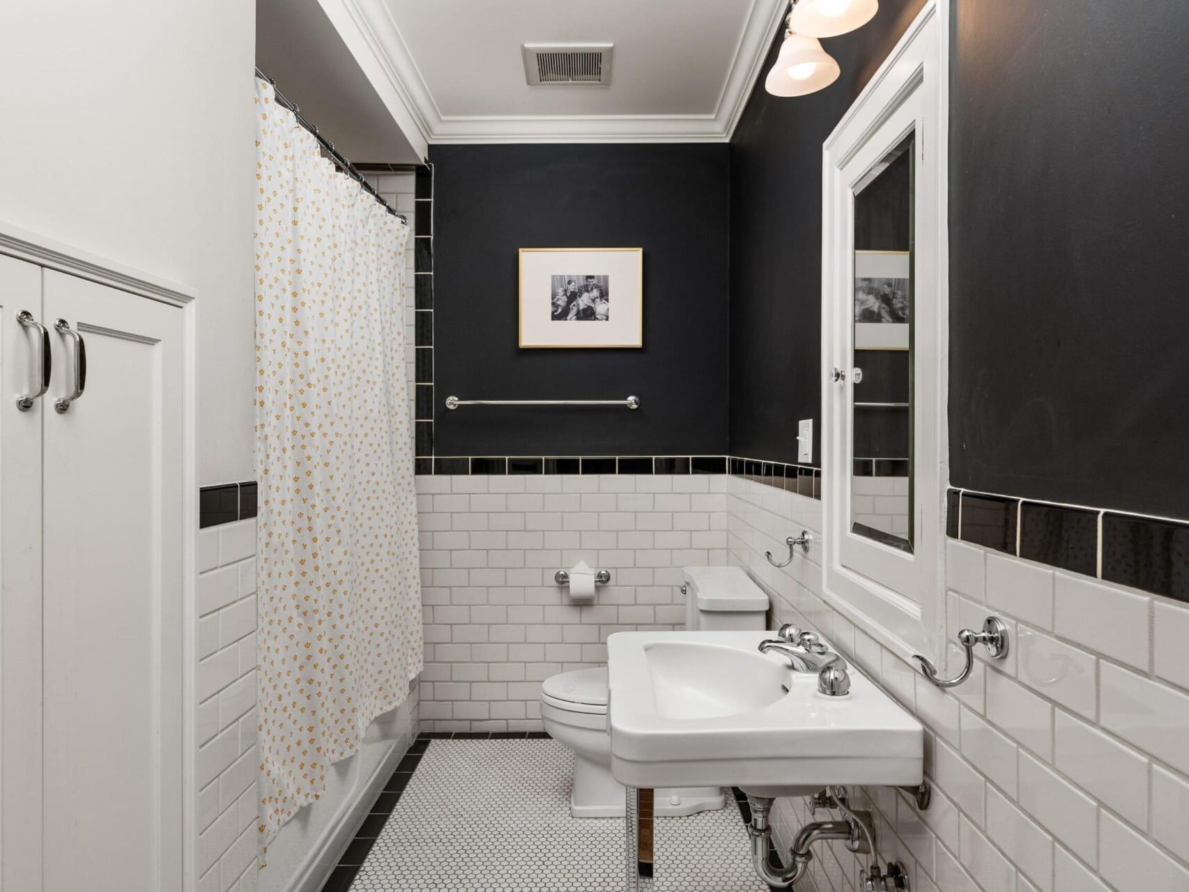 A bathroom in Portland, Oregon real estate showcases white tile walls with a black accent, a sleek white sink, toilet, and a bathtub adorned with a polka dot shower curtain. A mirror and framed picture grace the walls above the pristine white hexagonal tiled floor.