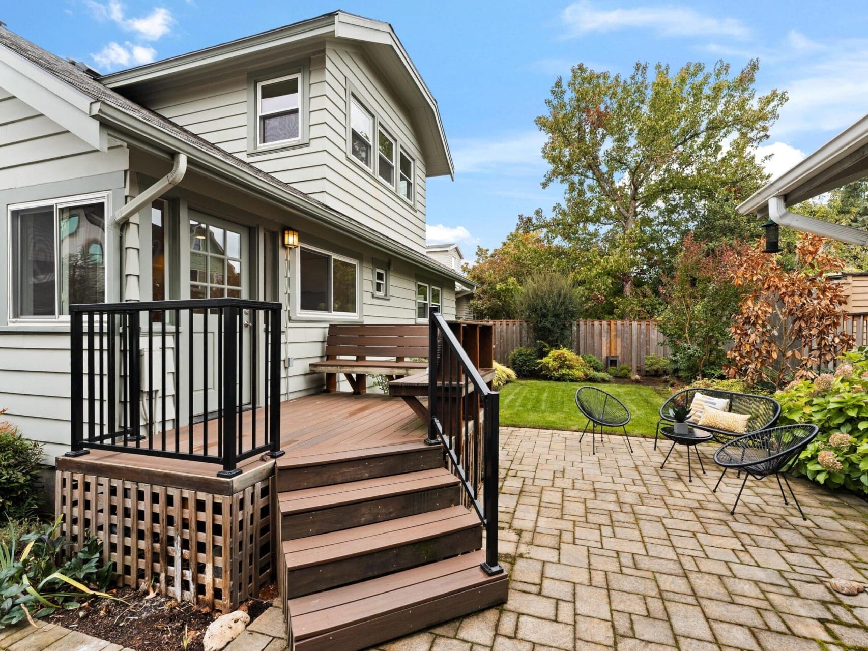 A cozy backyard with a wooden deck, steps with black railing, and bench seating. The patio features paver stones and modern metal chairs. The lawn is surrounded by greenery and trees, with a wooden fence in the background under a clear blue sky.