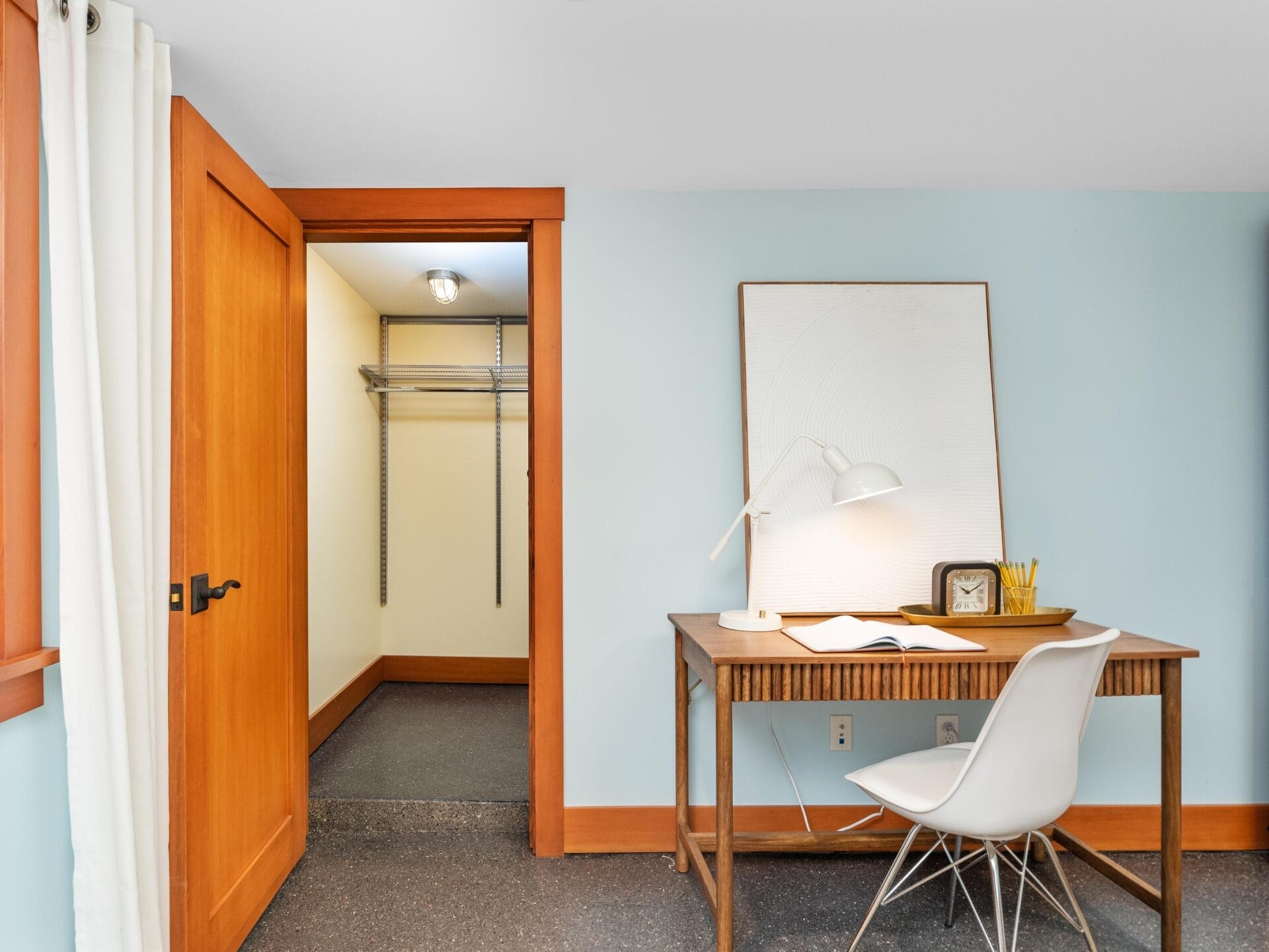 A minimalist study area featuring a wooden desk, a white chair, and a lamp captures the vibe of Portland, Oregon. An open doorway reveals a small walk-in closet with shelving. Light blue walls and wood trim give the space a warm, serene feel, reminiscent of Portland's laid-back charm.