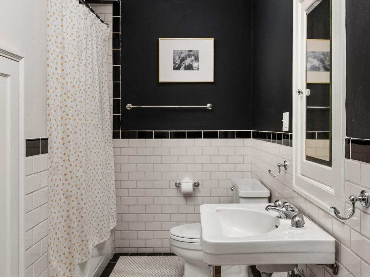 A small bathroom in a Portland, Oregon real estate gem features white subway tile walls, a black upper wall, and hexagonal floor tiles. There's a white sink, toilet, and shower with a polka dot curtain. A framed picture and light fixture are above the sink—ideal for showcasing by your Portland realtor.