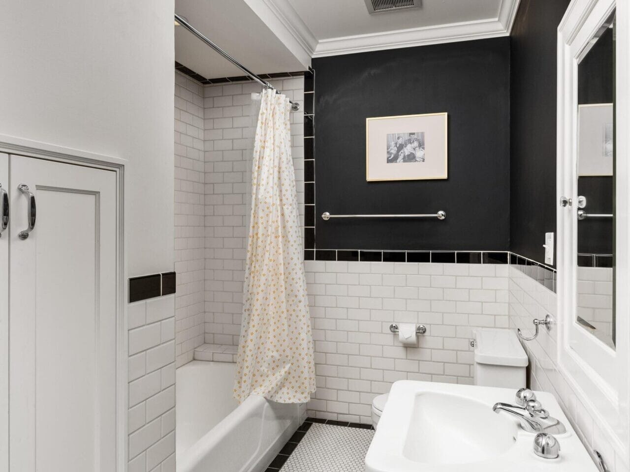 A small bathroom in a Portland Oregon Real Estate listing features white subway tiles, a combination bathtub and shower with a white and beige curtain, and black accent tiles. The room showcases a white sink, light fixture above the mirror, and cabinet, with white hexagonal floor tiles.