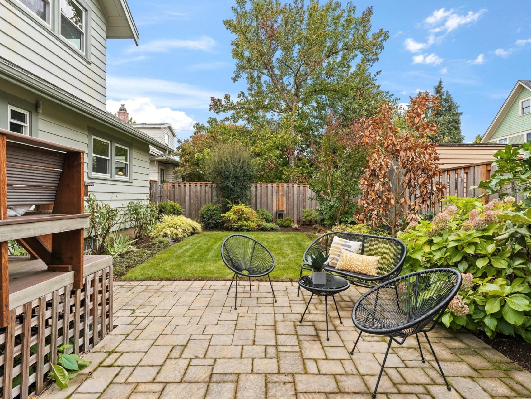 A backyard patio featuring three black wire chairs and a round table on a brick surface. The lawn is bordered by a variety of green shrubs and trees. A wooden fence surrounds the yard, and a gray house is visible on the left. The sky is blue with clouds.