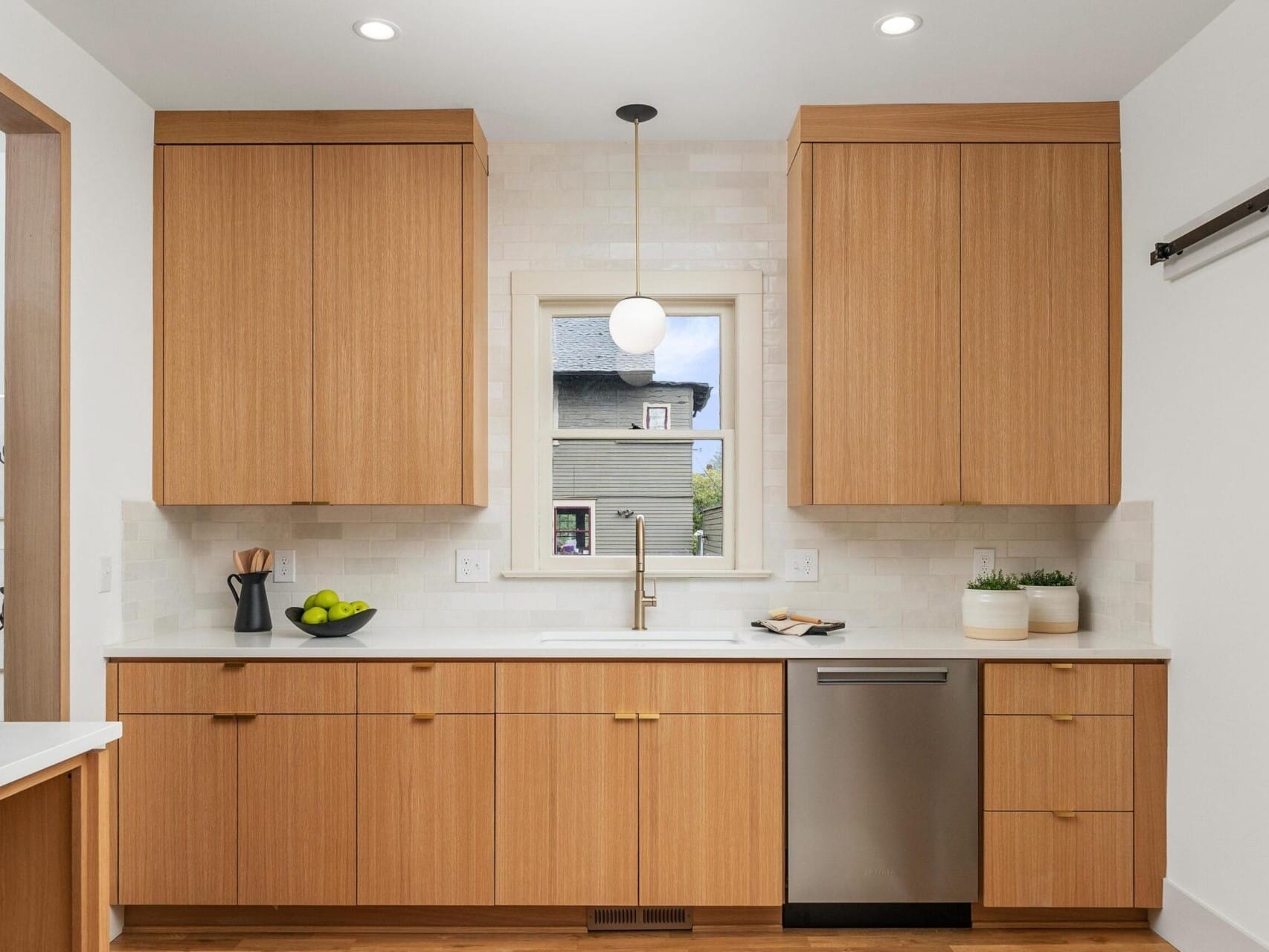 A modern kitchen in a chic Portland, Oregon style features wooden cabinets and white countertops. Natural light pours through a central window above the sink. A pendant light hangs overhead, while a stainless steel dishwasher sits to the right and a fruit bowl with green apples adorns the counter.