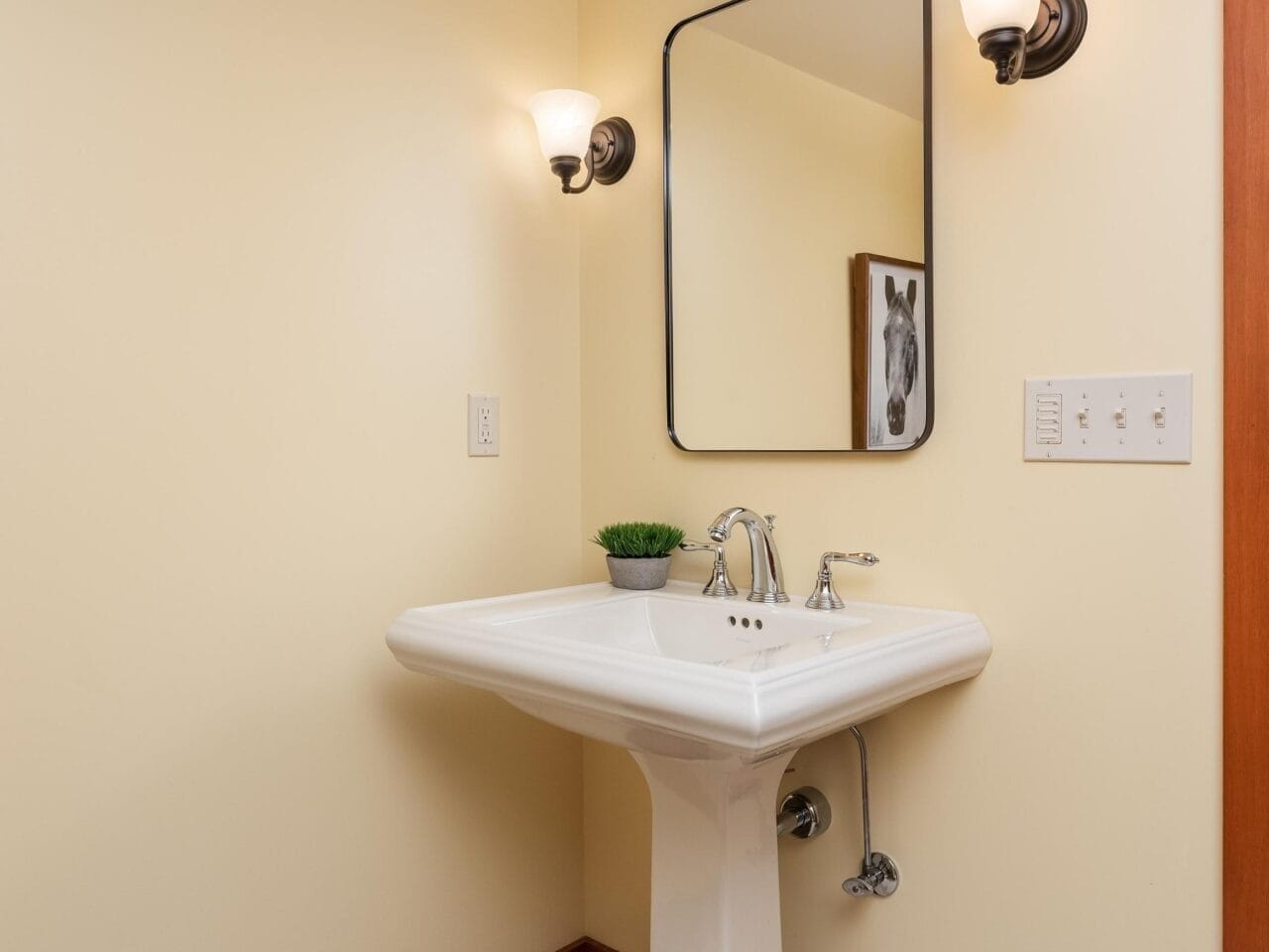 A bathroom corner in Portland, Oregon, with pale yellow walls features a white pedestal sink, a rectangular mirror above, and two wall-mounted sconces. A small plant sits on the sink, and a light switch is visible on the wall.