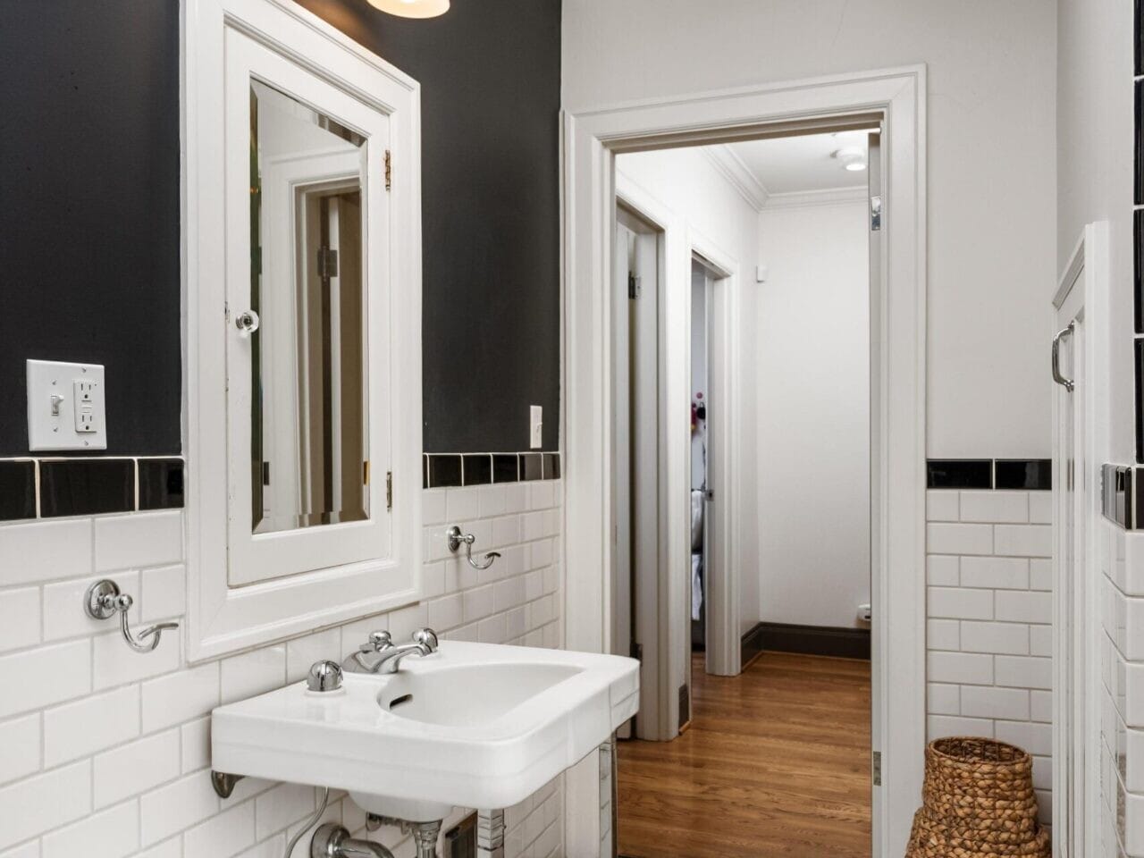 A bathroom with a white sink and chrome fixtures beneath a wall-mounted mirror. Black and white subway tiles line the walls, while hexagonal tiles cover the floor. A wicker basket stands in the corner, complementing this Portland Realtor's elegant design, with a hallway visible through an open door.