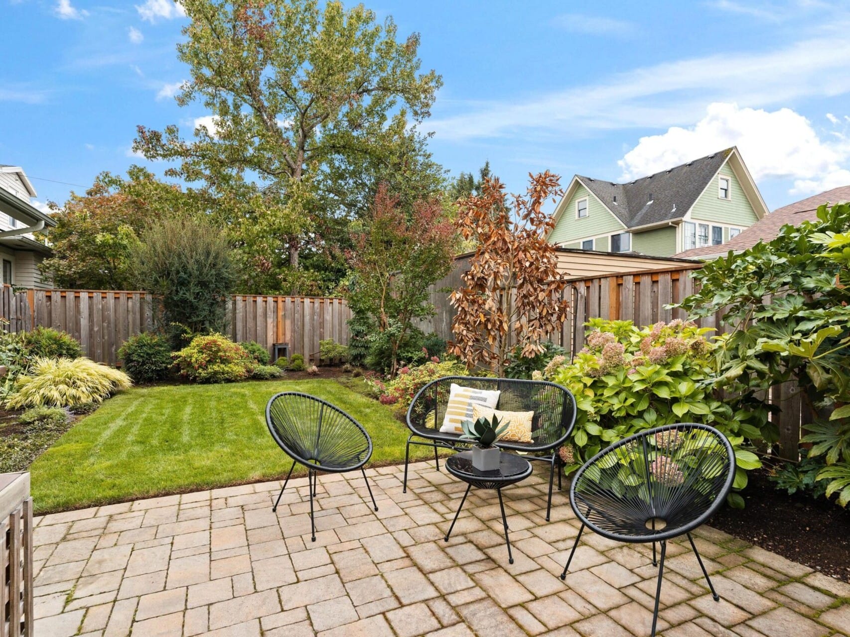 A backyard features a paved patio with three black chairs around a small table. Lush green grass and various shrubs are visible, surrounded by a wooden fence. In the background, there are trees and a house under a clear blue sky.