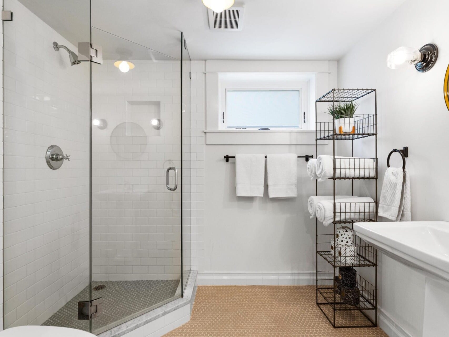 A modern bathroom in Portland, Oregon, features a glass-enclosed shower, white tiles, a pedestal sink, and a circular mirror. Towels are neatly arranged on a metal rack, while a small potted plant adds a touch of greenery to the space.