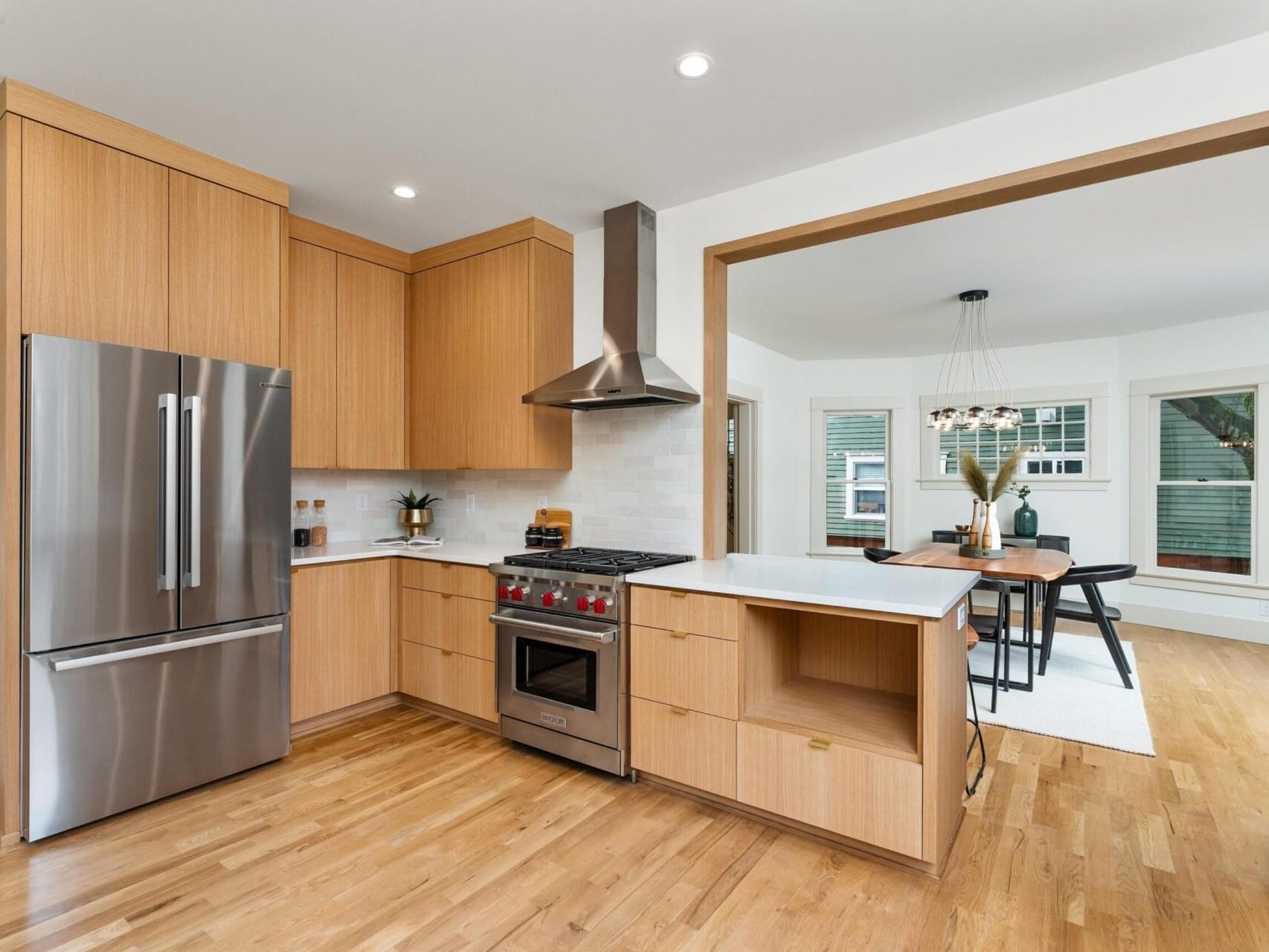 Located in Portland, Oregon, this modern kitchen features wooden cabinets, stainless steel appliances, and an island. It opens into a dining area with a dark table and chairs. Large windows flood the space with natural light, while a chandelier graces the dining table. Hardwood floors run throughout.