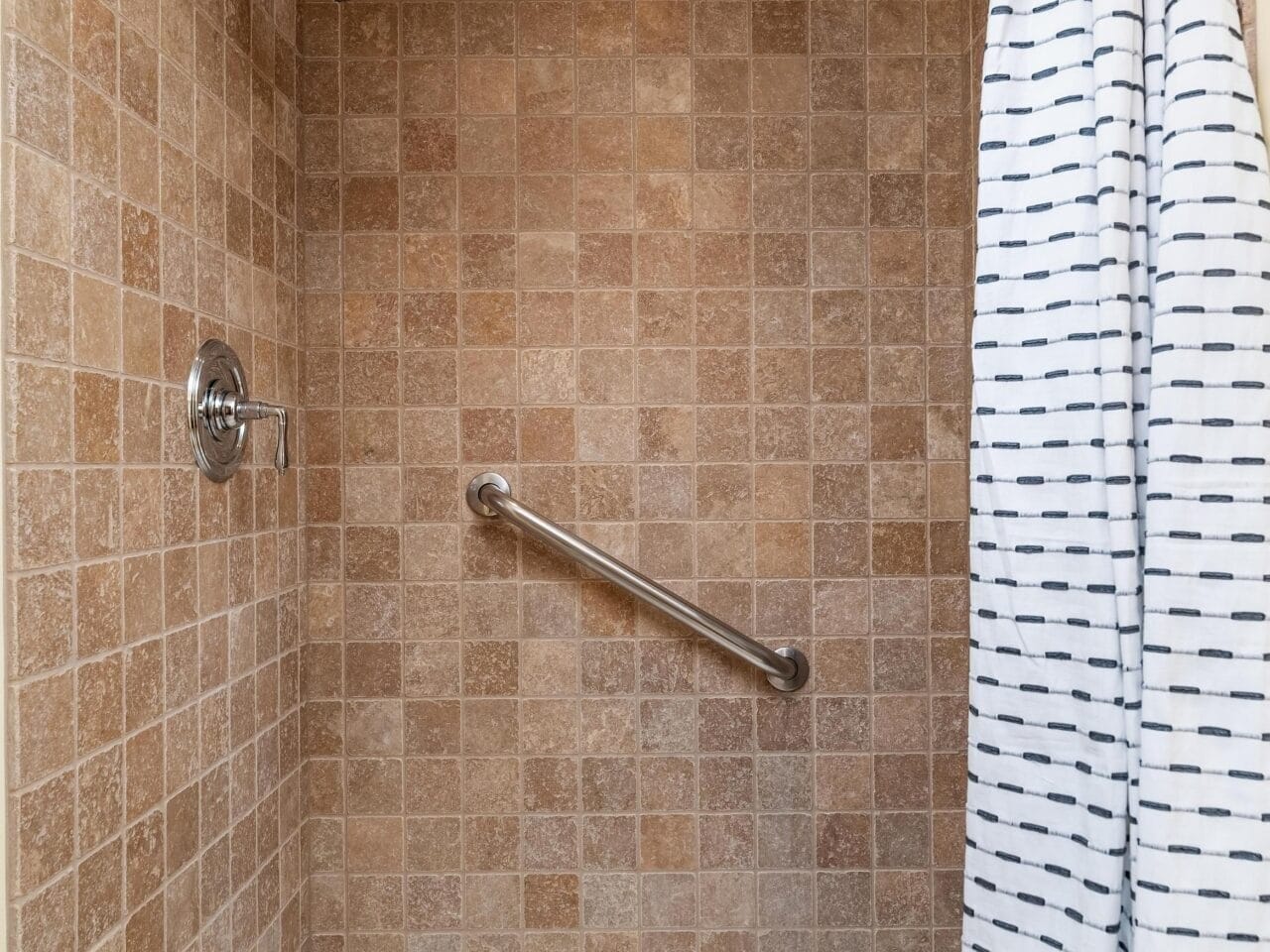 A shower in Portland, Oregon, features beige square tiles and a sleek stainless steel showerhead with an accompanying grab bar. A white curtain with horizontal black stripes is partially drawn across the entrance, while the floor showcases a white base with a central drain.